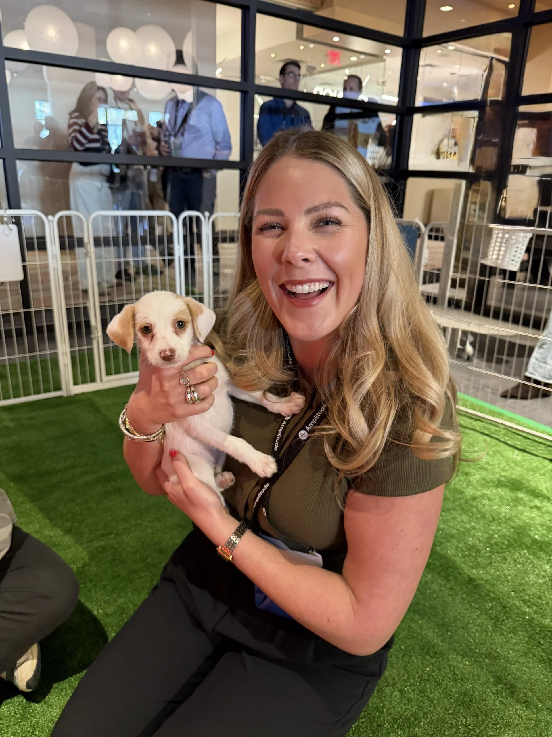 Smiling woman with blonde hair kneeling on artificial turf holding a small light-colored puppy