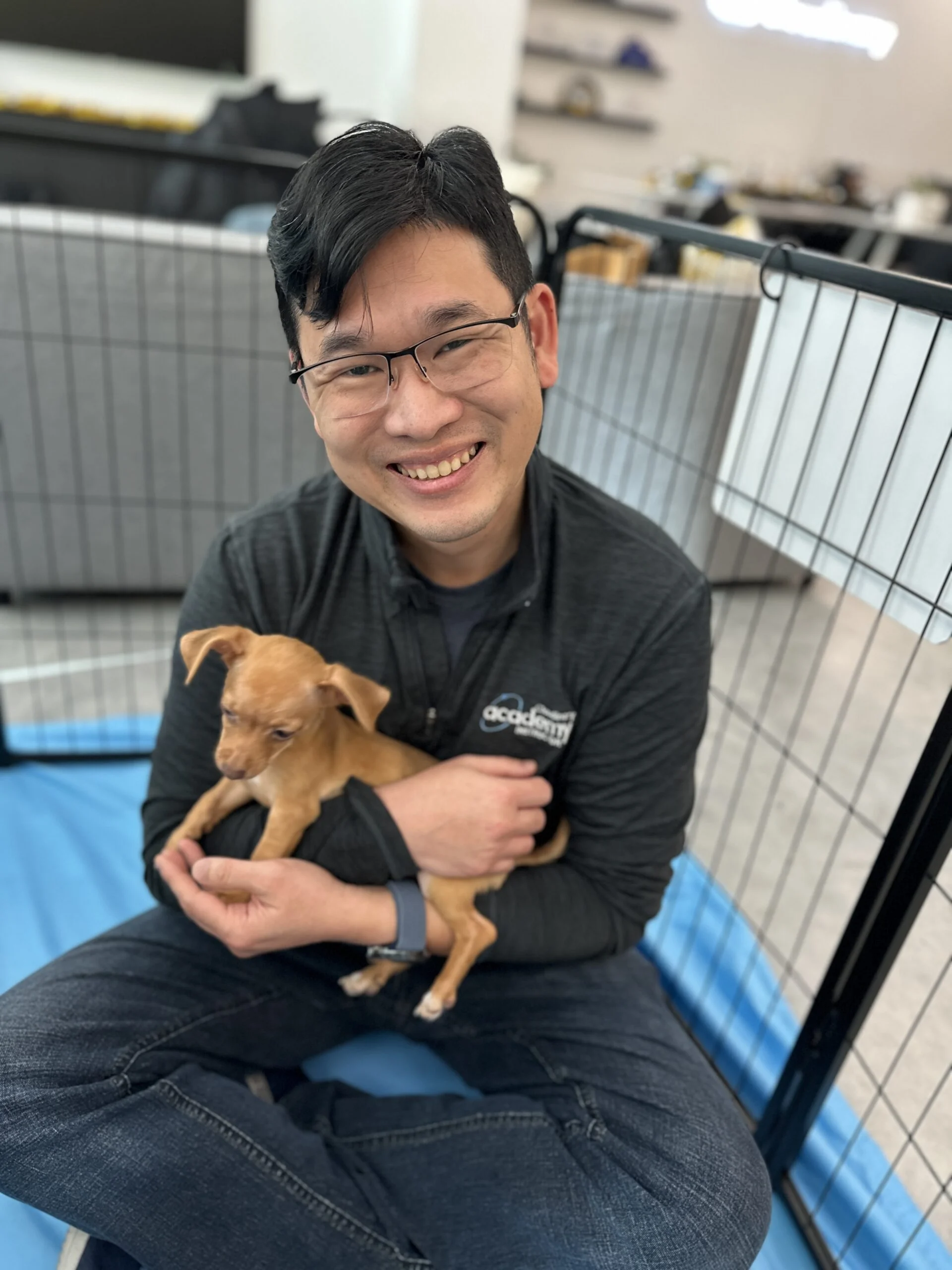Smiling man with glasses sitting indoors holding a small tan puppy on a blue floor mat