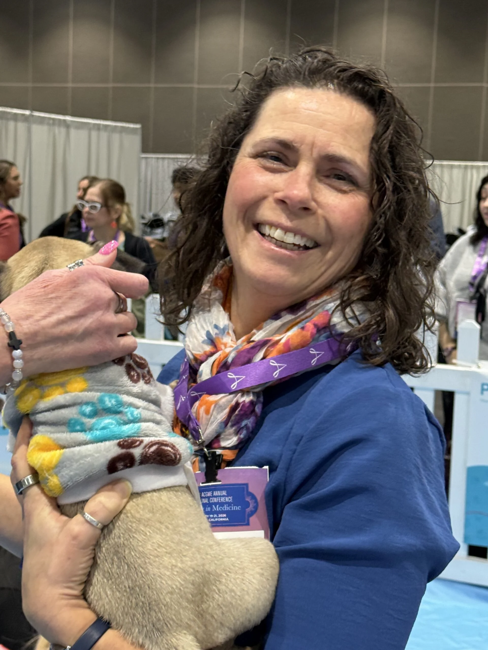 Smiling woman wearing a blue jacket and colorful scarf holding a small puppy wrapped in a sweater