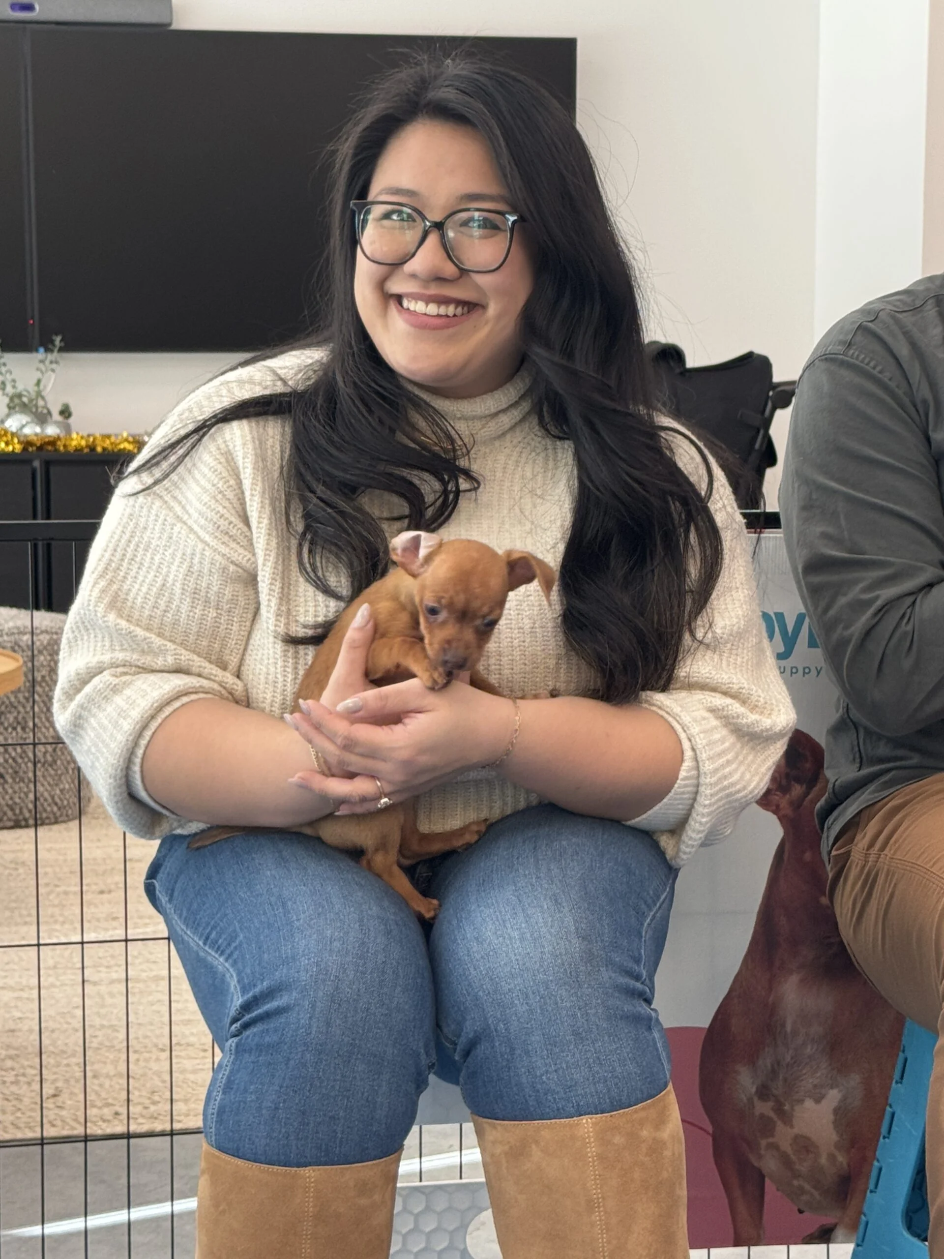 Smiling woman wearing glasses and a cream sweater sitting indoors while holding a small brown puppy
