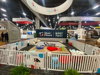 A wide-angle shot of an empty puppy play area featuring wood-look flooring enclosed by a white picket fence, with a large backdrop reading "shaw floors | pet perfect. "