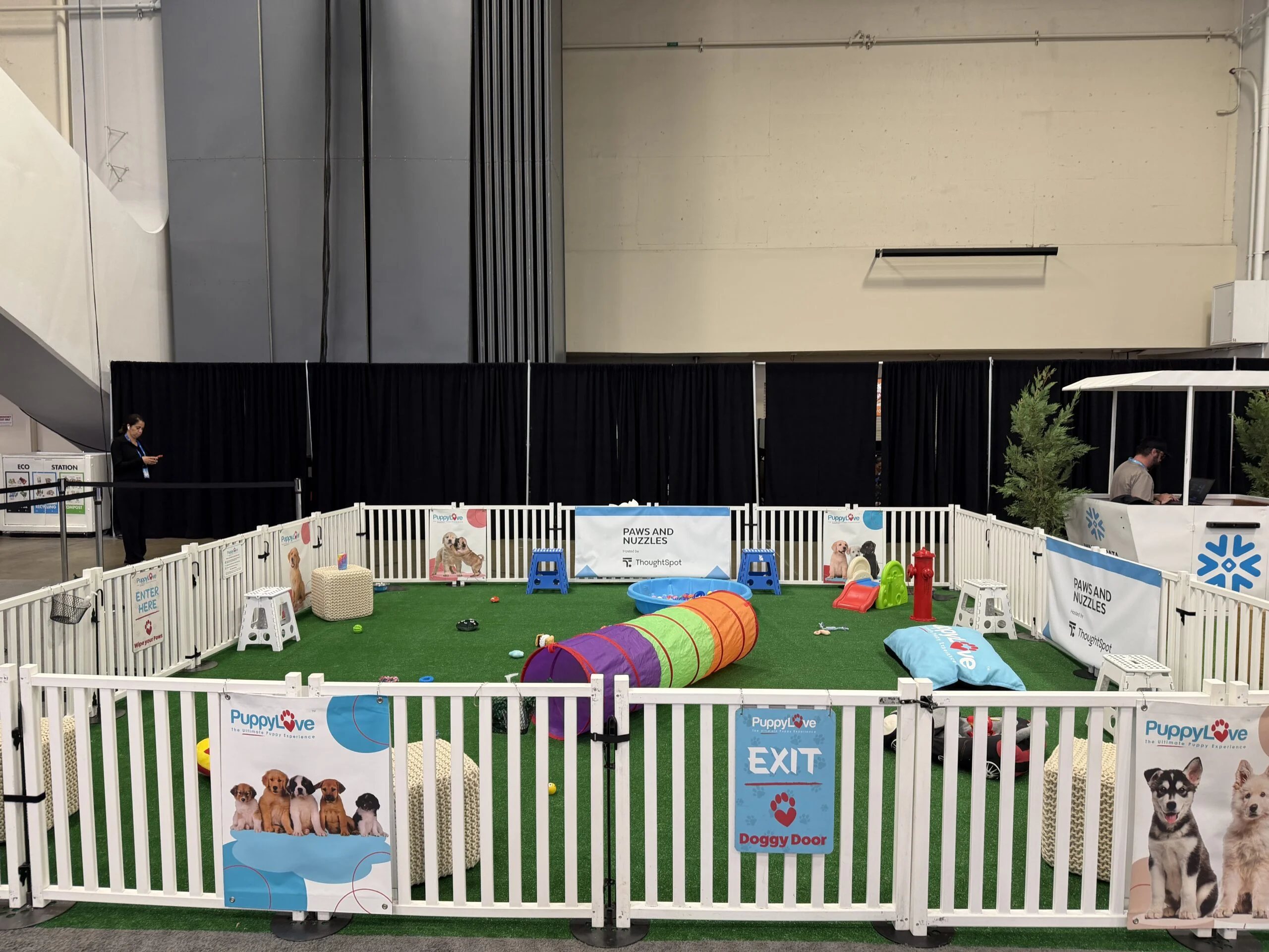 An empty puppy play area with artificial green turf enclosed by a white picket fence, featuring a colorful crawl tunnel, a blue plastic pool, and signs reading "paws and nuzzles" and "puppylove. "