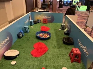 An overhead view of an empty puppy play area with artificial green turf enclosed by light blue panels reading "safesend" and "puppylove," featuring a central colorful ball pit, red heart-shaped rugs, dog beds, and plastic stools.