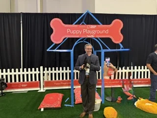 A smiling man in a blazer stands holding a puppy under a blue dog-house-shaped frame with a large red bone sign reading "puppy playground" while someone takes a photo of him.