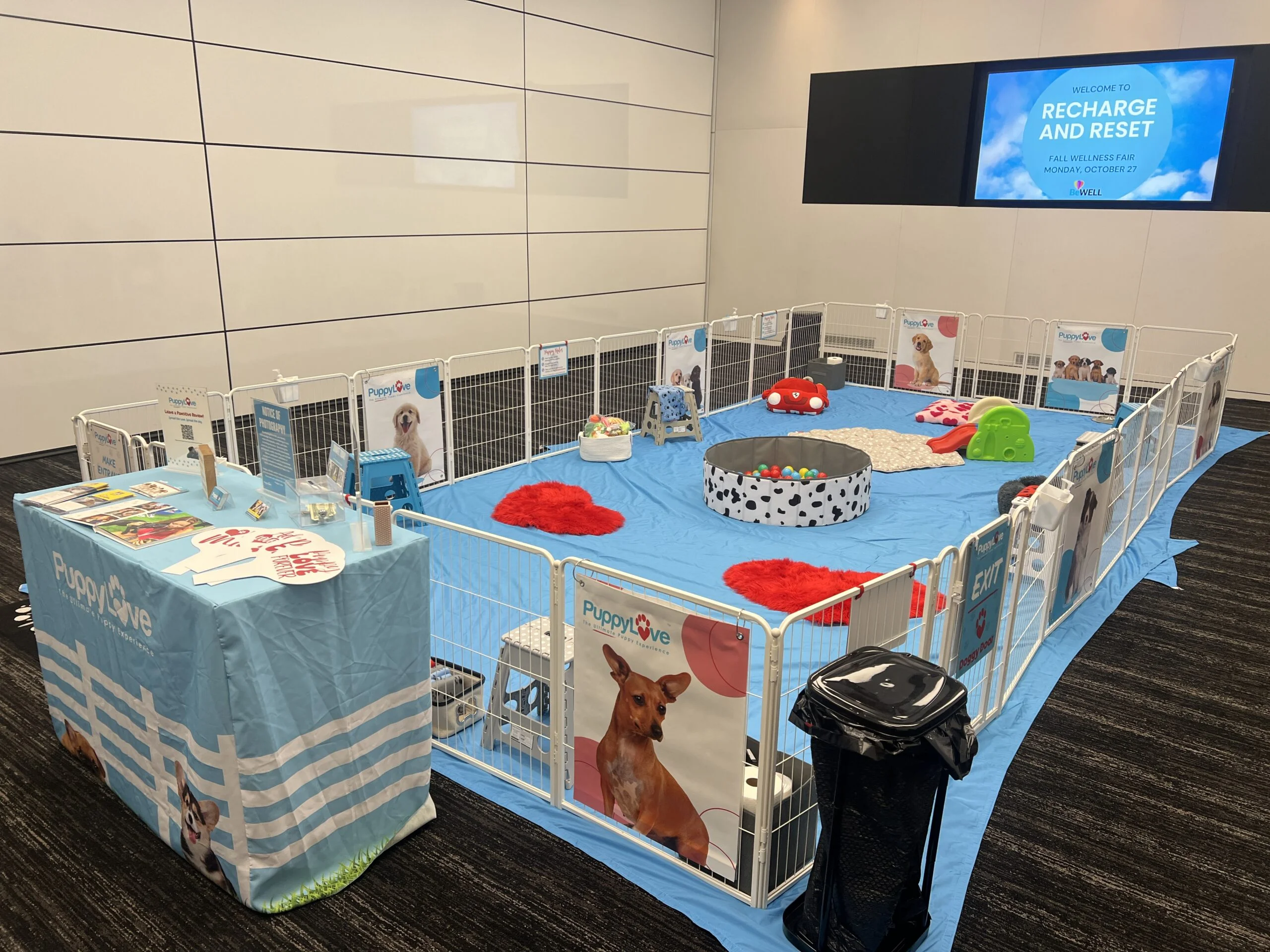 An empty puppy play area with a light blue floor tarp enclosed by a white wire fence, featuring a dalmatian-print ball pit, a mini slide, red heart rugs, and a branded puppylove welcome table beneath a screen reading "recharge and reset. "