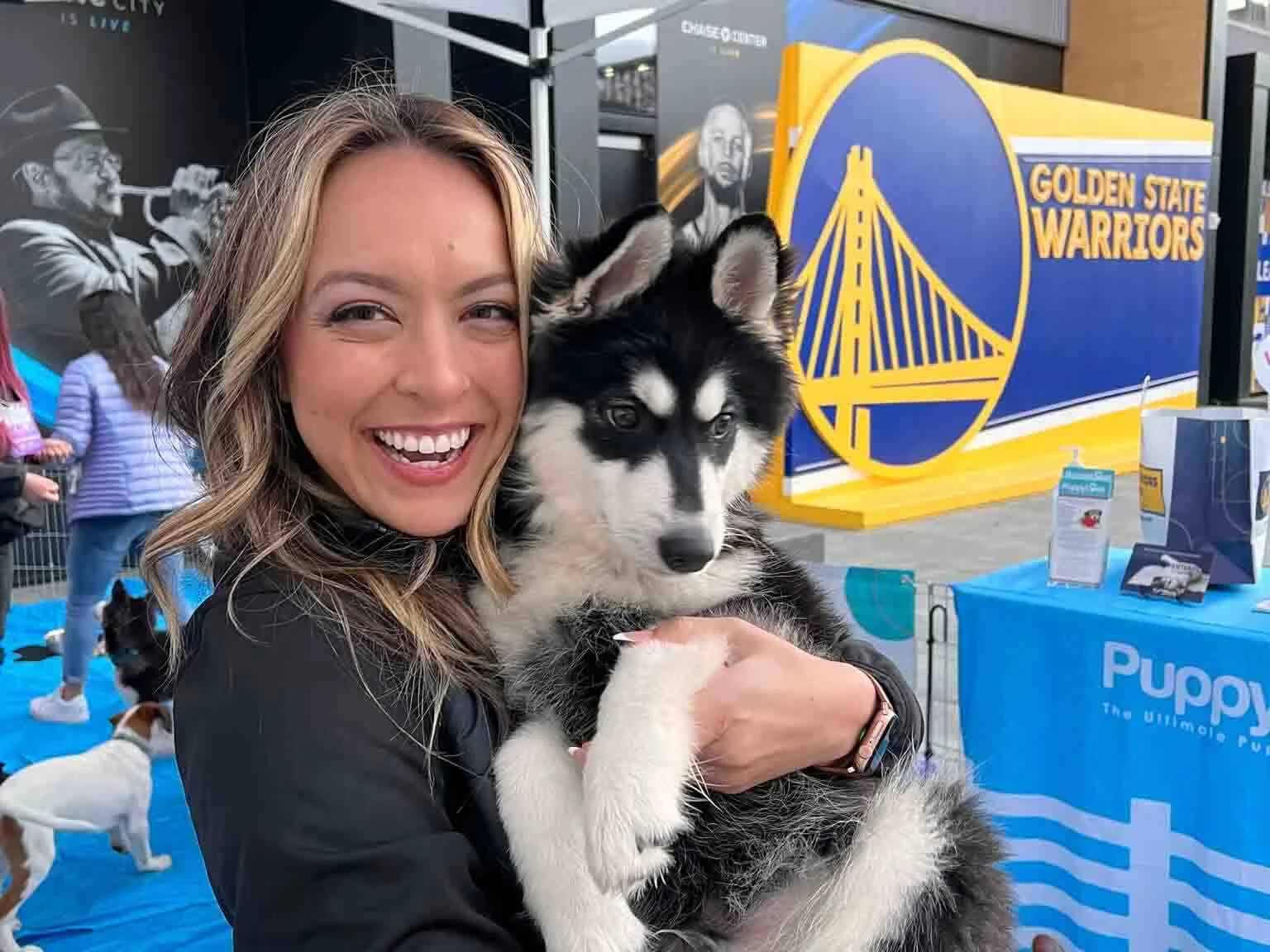 A woman holding a black and white husky puppy in front of a golden state warriors banner