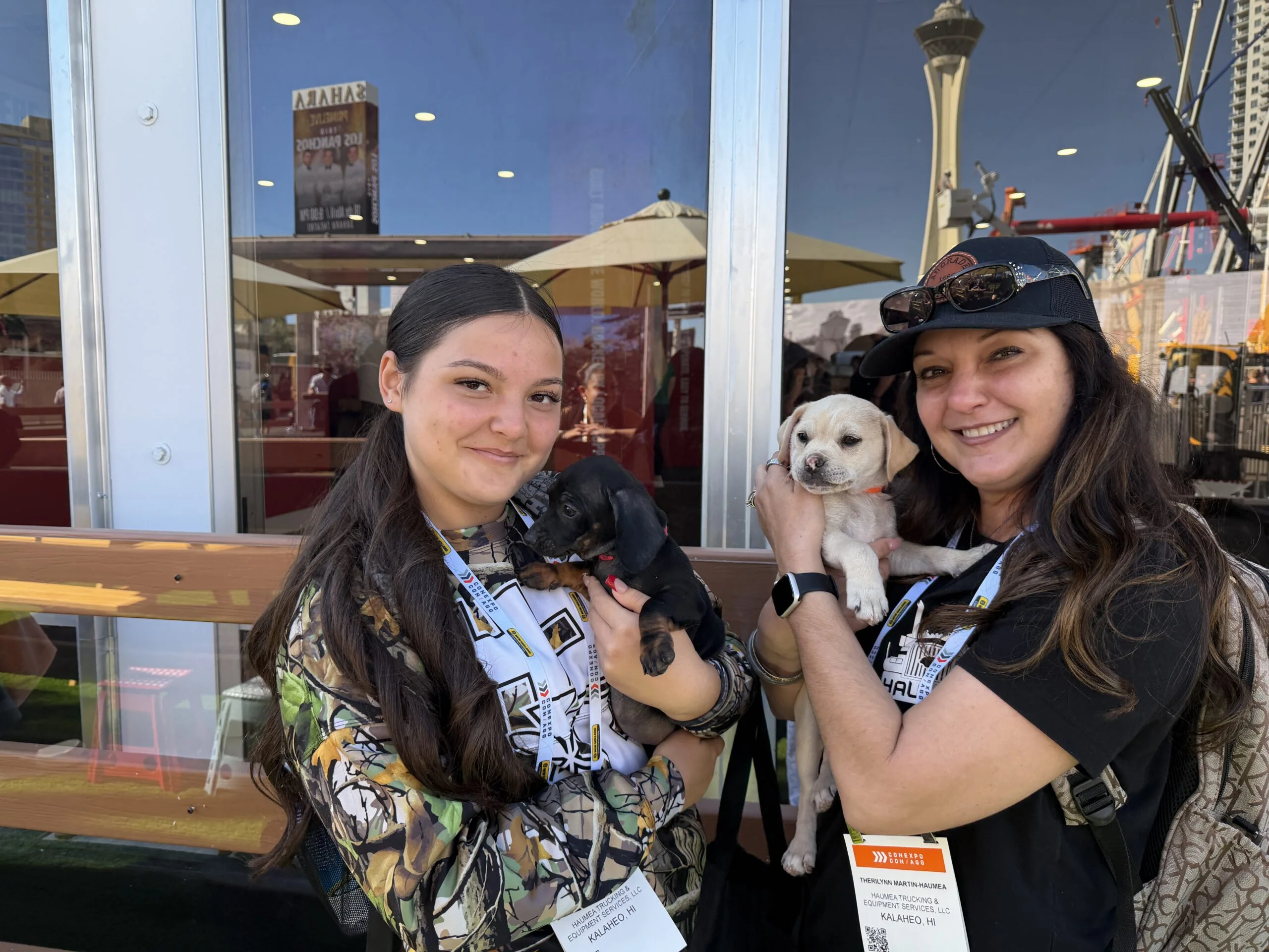 Two women smiling outdoors while holding a small black puppy and a small cream puppy