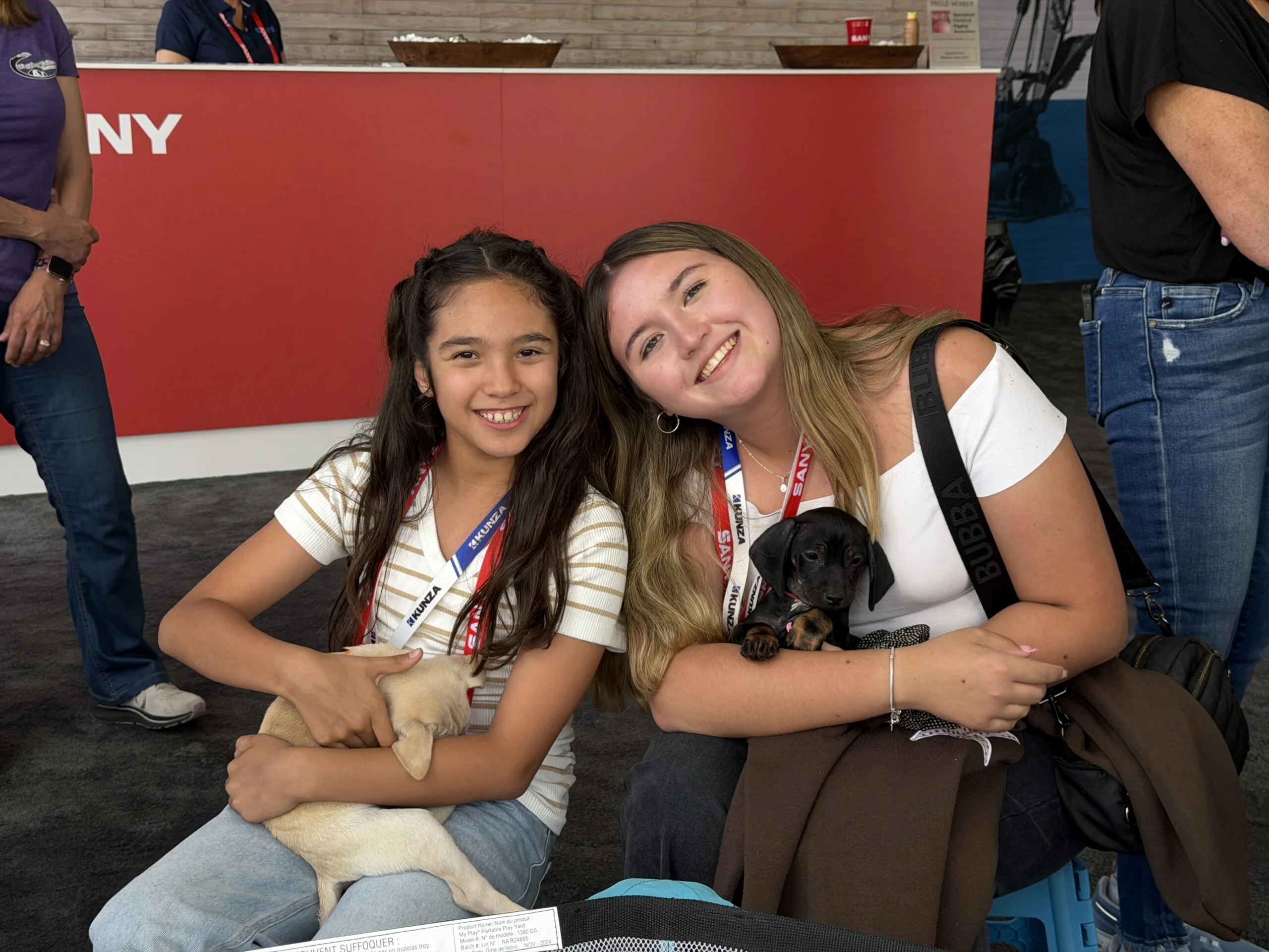 Two young women sitting together holding small puppies at an indoor event