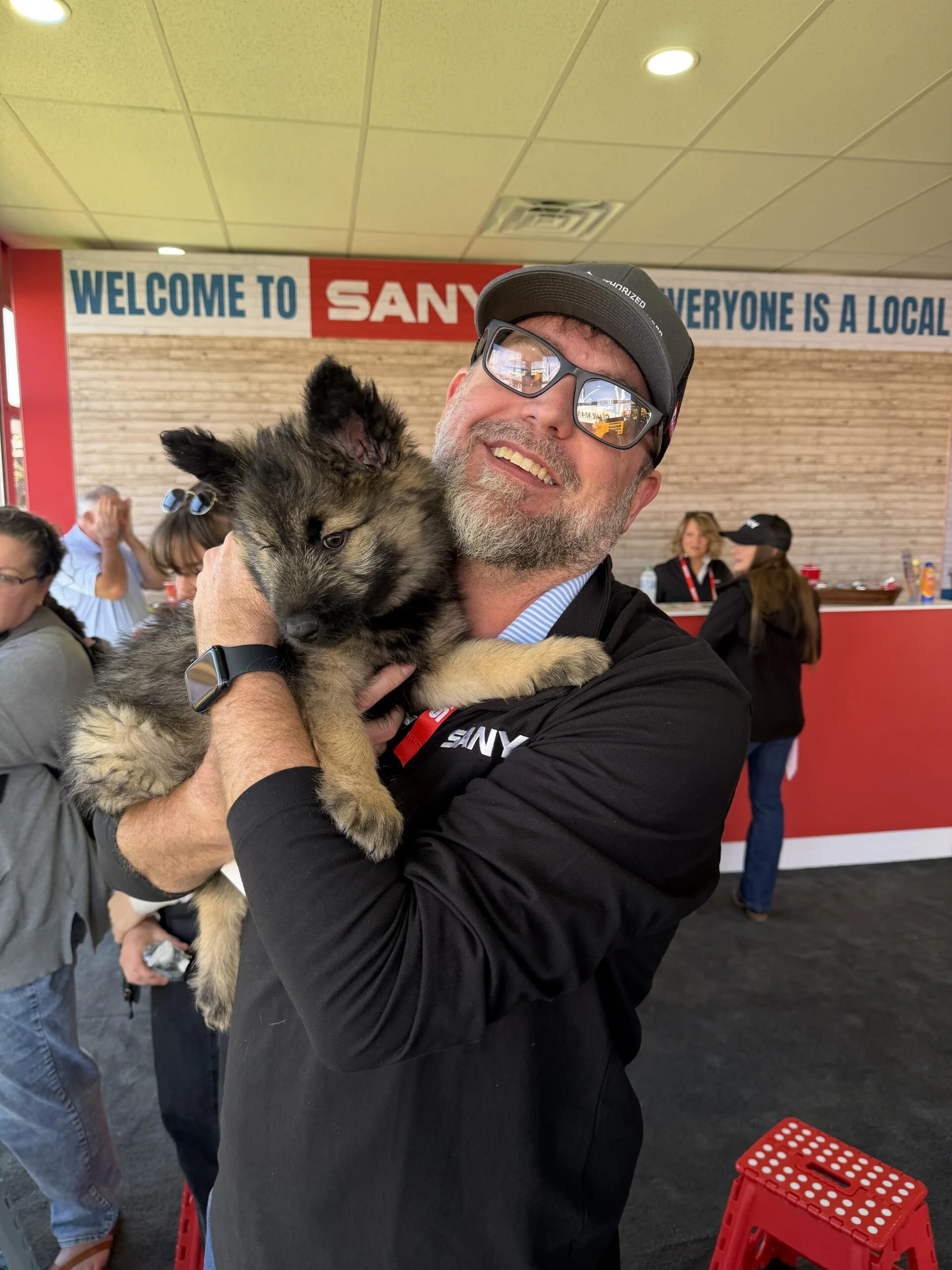 Smiling man in a black sany shirt and hat holding a fluffy german shepherd puppy indoors