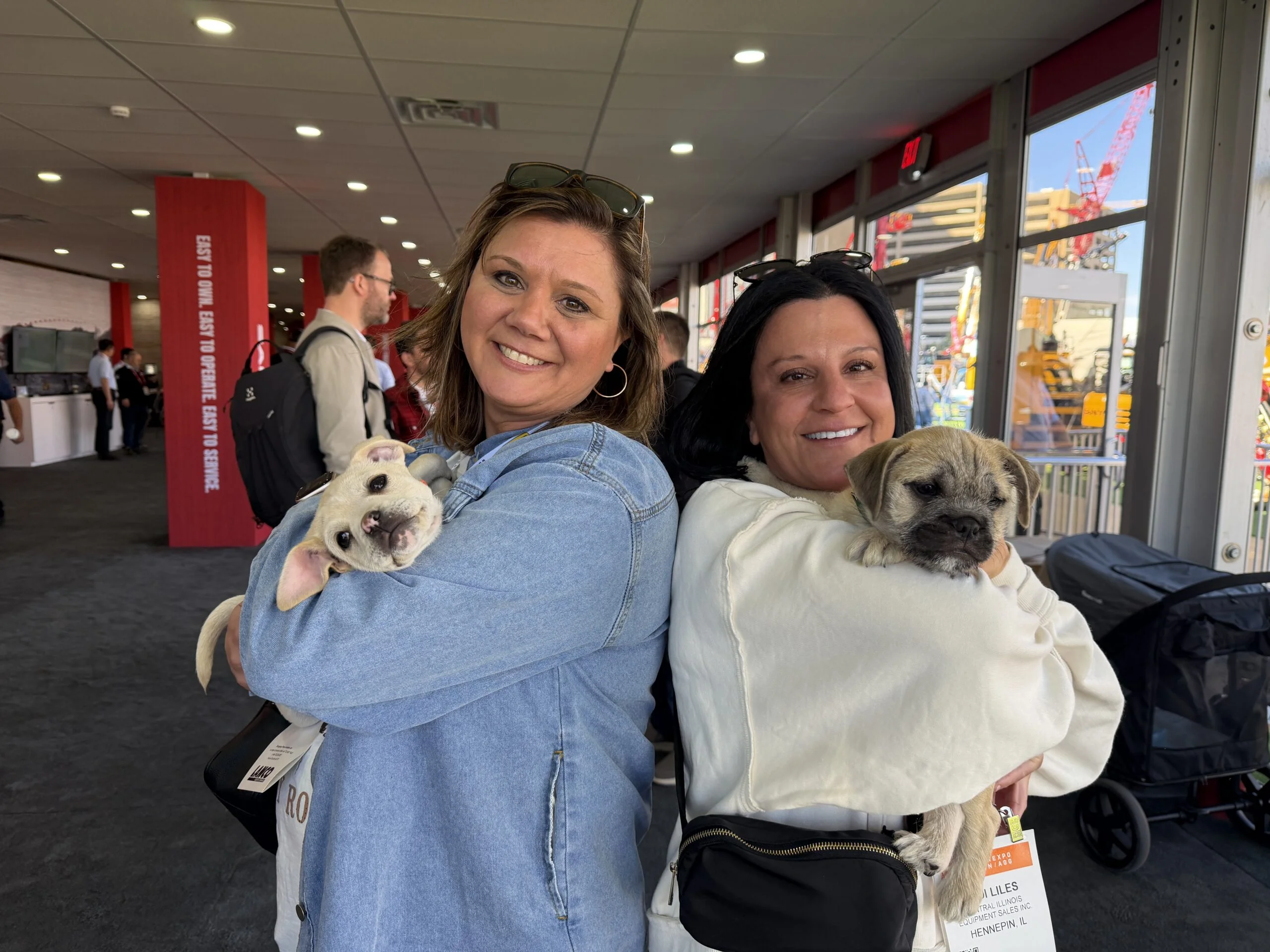 Two women smiling indoors while each holds a small puppy at an event