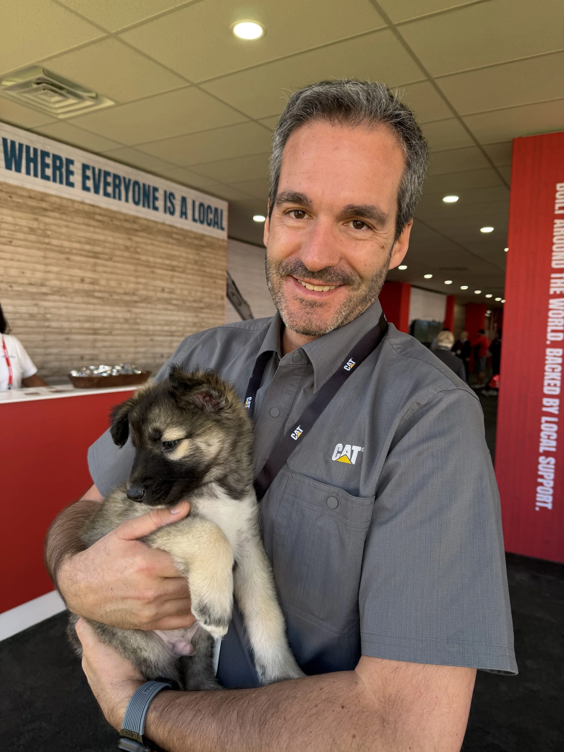 Smiling man in a gray cat shirt holding a small brown puppy indoors
