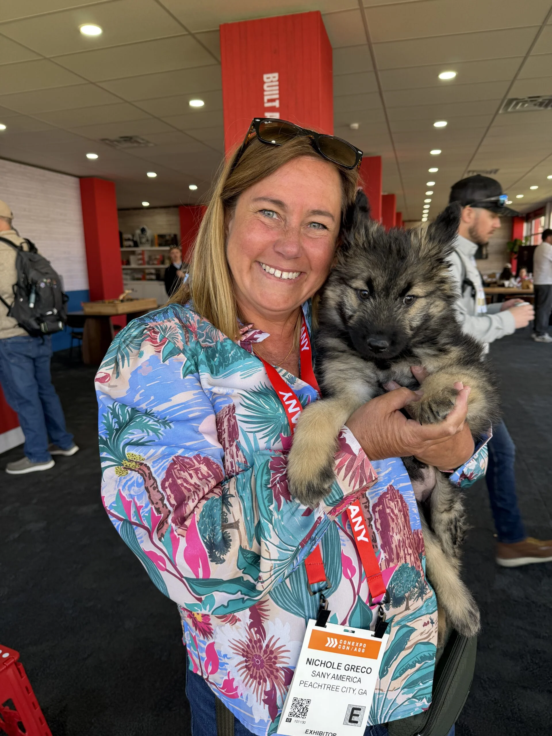 Woman in a colorful shirt with sunglasses on her head holding a fluffy gray and black puppy