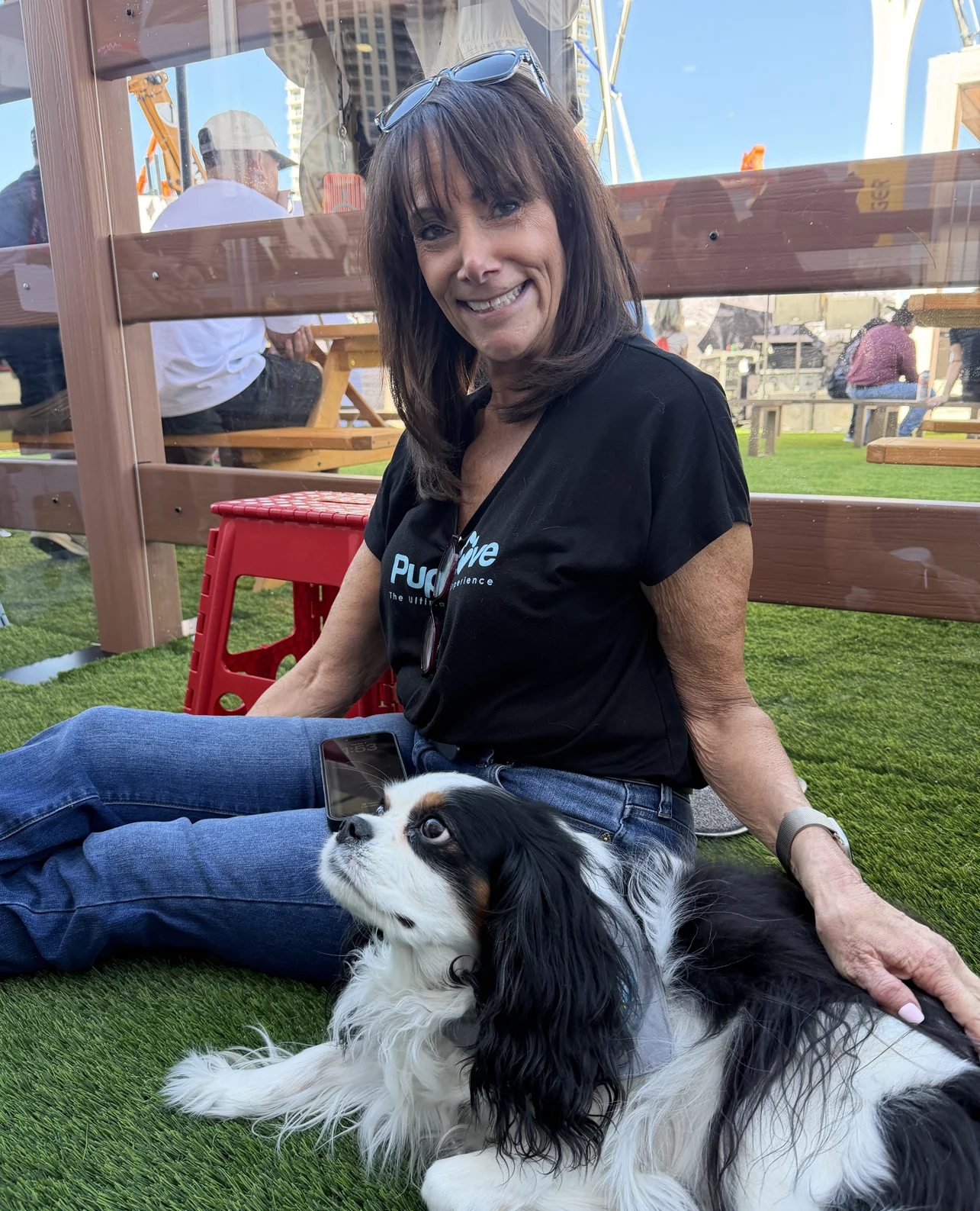 Smiling woman with sunglasses on her head sitting outdoors on artificial grass next to a black and white dog