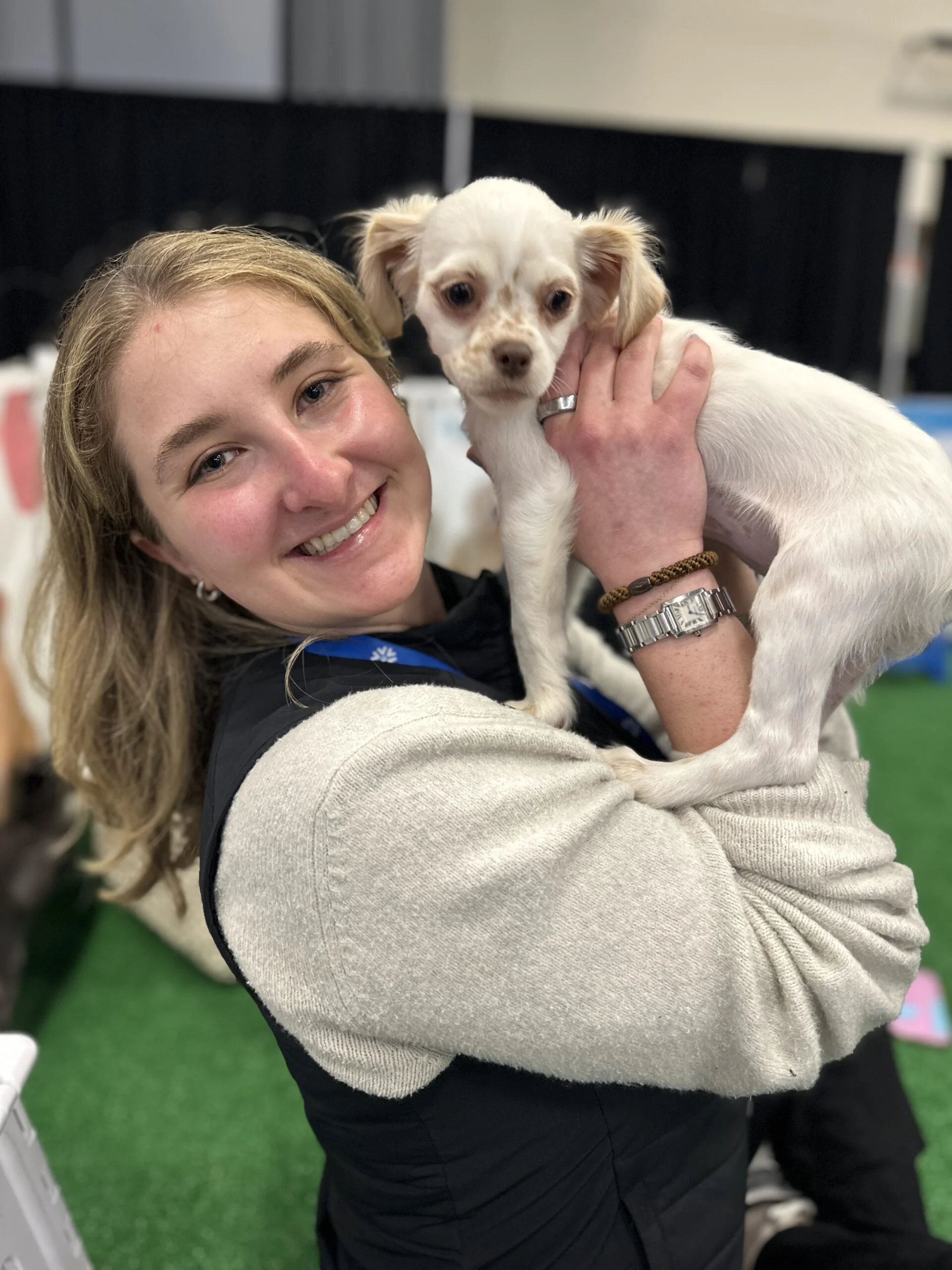A smiling woman with light brown hair, wearing a beige sweater sleeve under a black vest, holds a small white puppy with floppy ears up in her hands indoors.