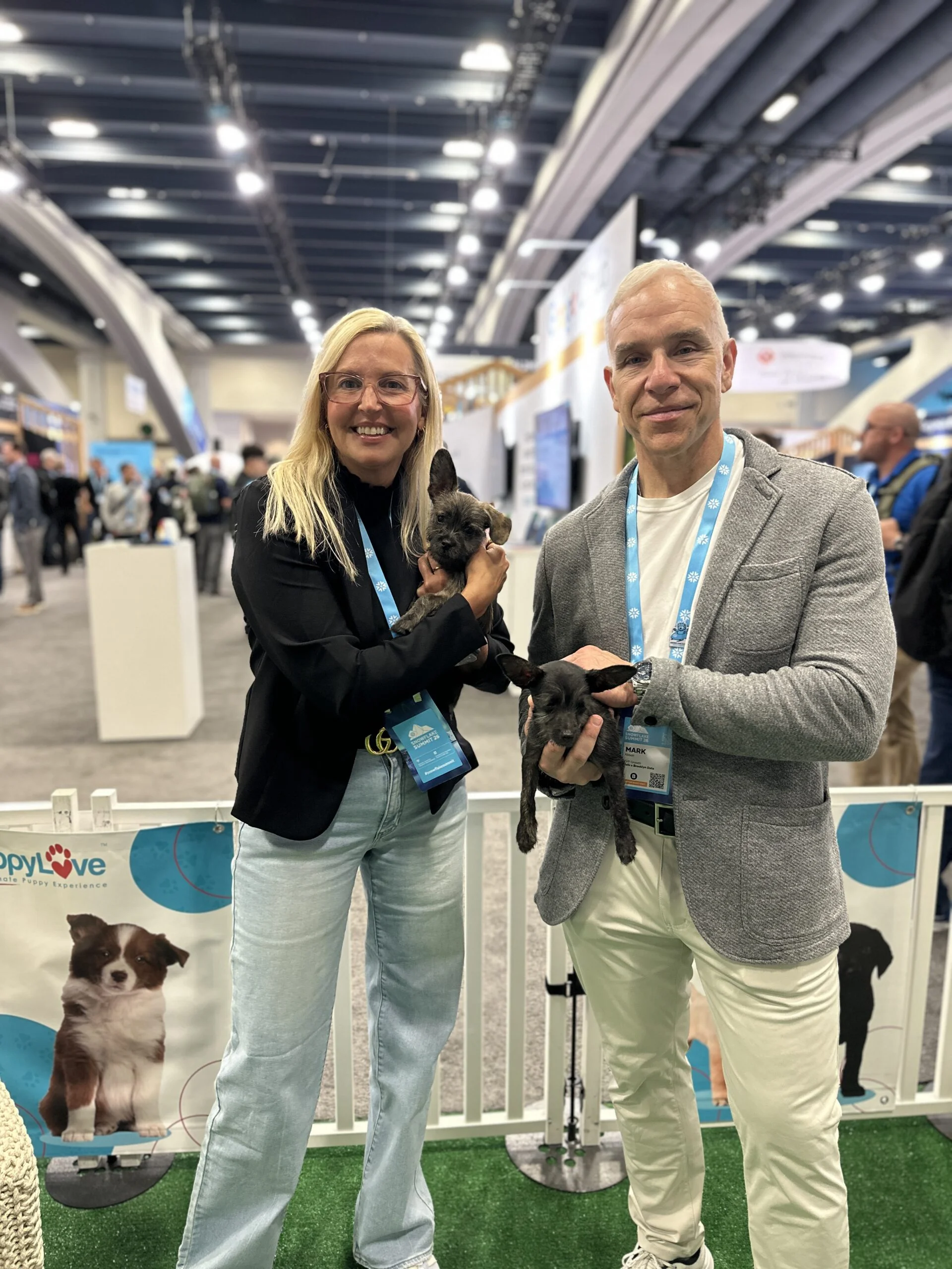 A blonde woman in a black blazer and light jeans stands next to a man with short gray hair in a gray jacket, with each person holding a small puppy at an event.