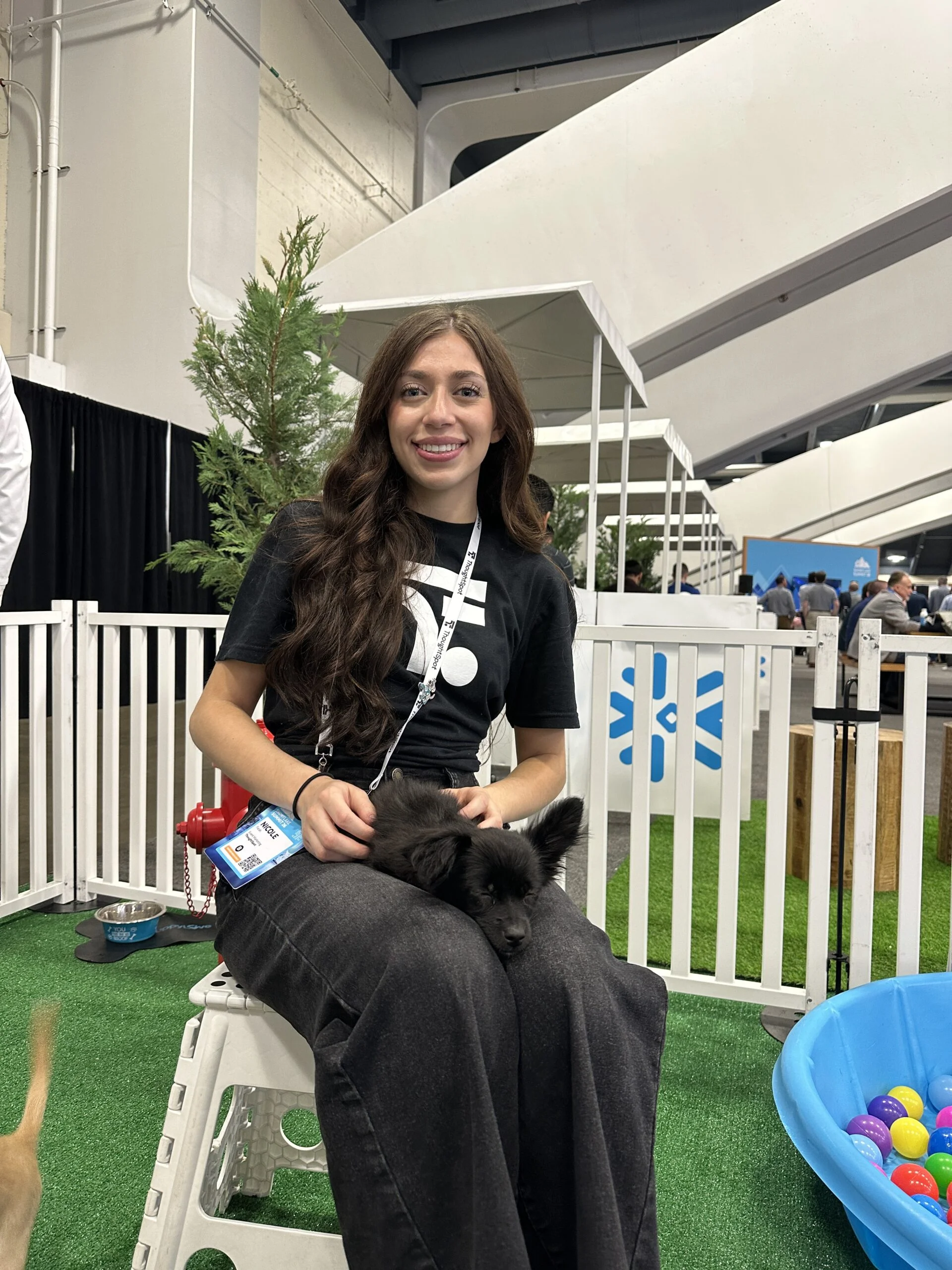 A smiling woman named nicole with long brown hair, wearing a black graphic t-shirt and dark jeans, sits on a white plastic stool while a small black puppy rests comfortably in her lap.