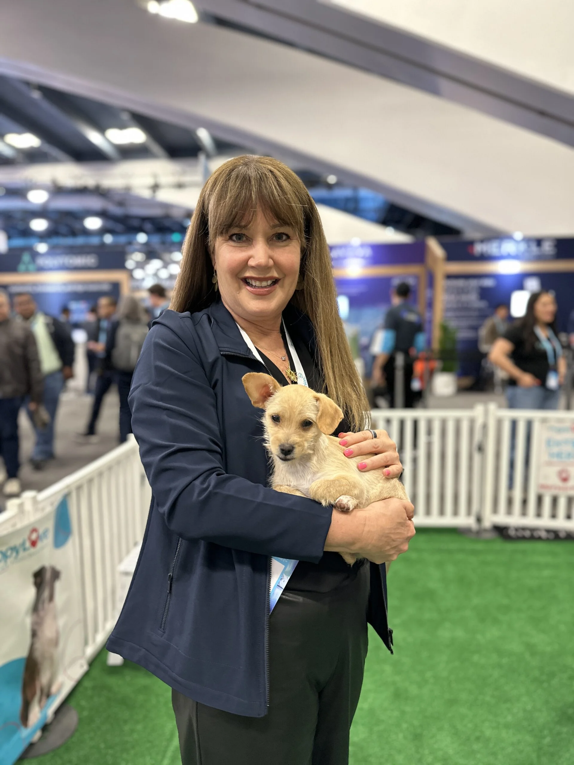 A smiling woman with straight brown hair and bangs, wearing a dark navy blue jacket and dark trousers, holds a small wire-haired light tan puppy in her arms.