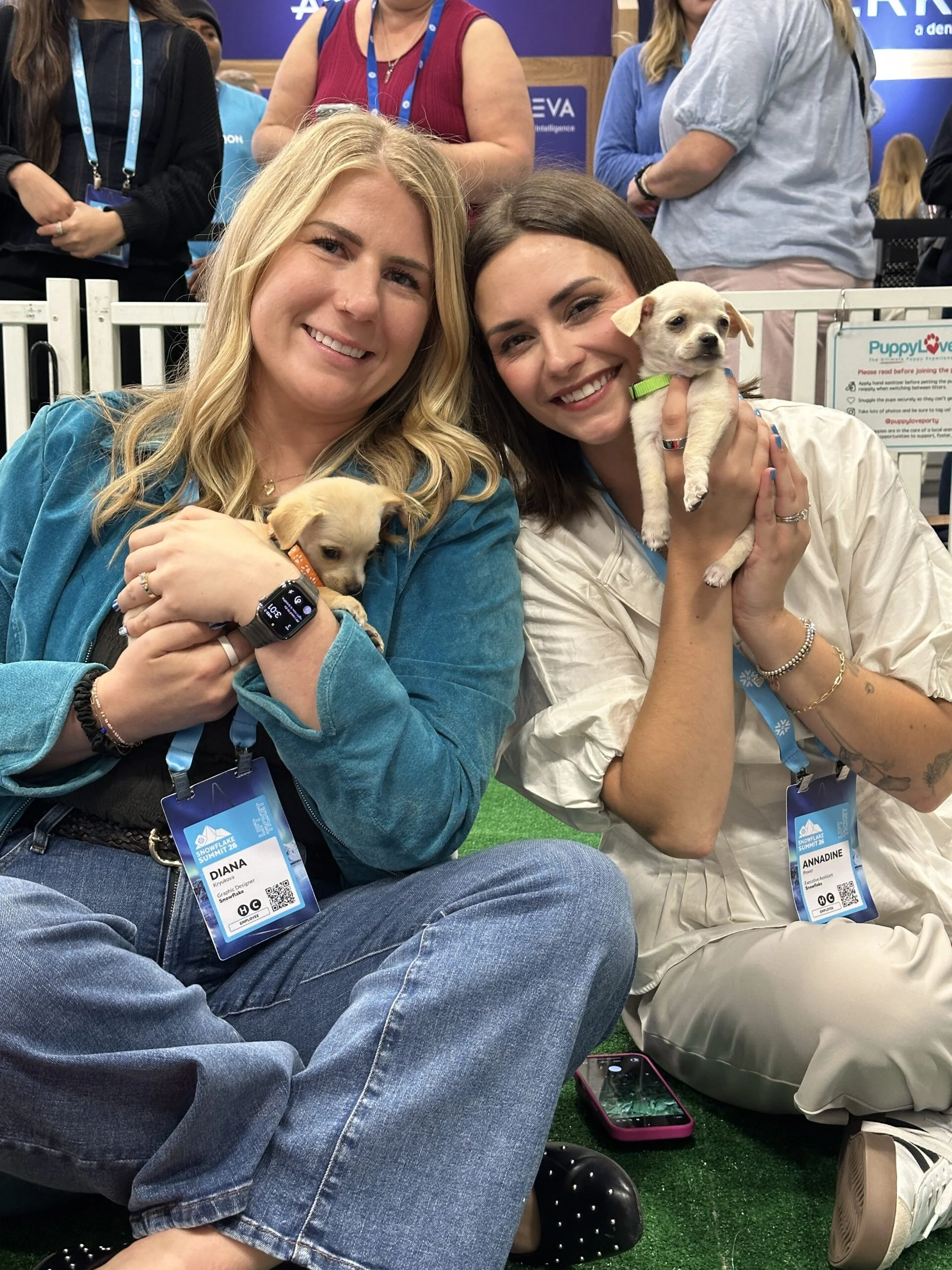 Two smiling women, named diana and annadine, sit side-by-side on artificial turf, each holding a tiny cream-colored puppy in their hands.