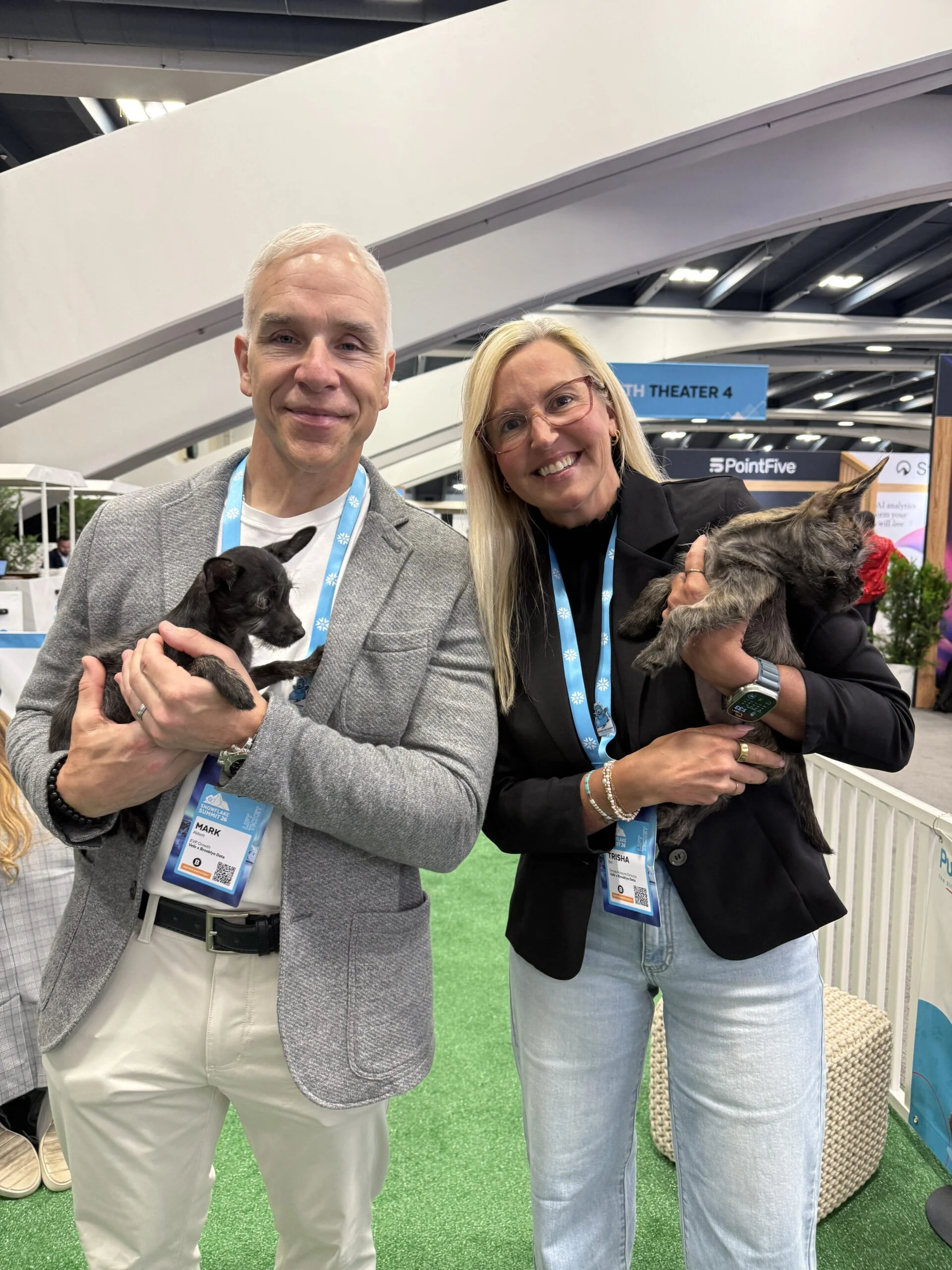 A man named mark with short gray hair in a gray jacket and a blonde woman named trisha in a black blazer stand together indoors, each holding a small puppy in their arms.