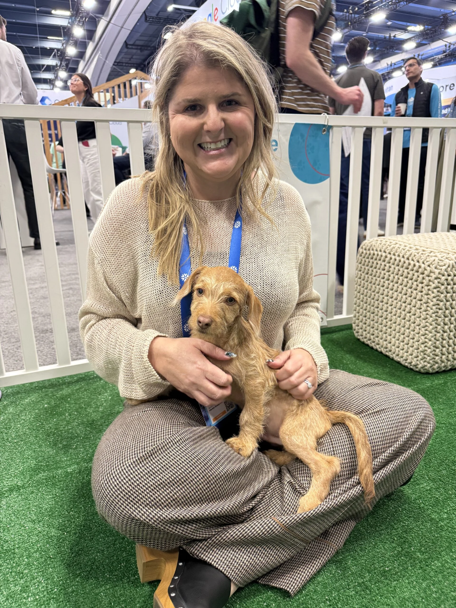 A smiling blonde woman wearing a light beige loose-knit sweater and pattern trousers sits cross-legged on artificial turf holding a wire-haired tan puppy.