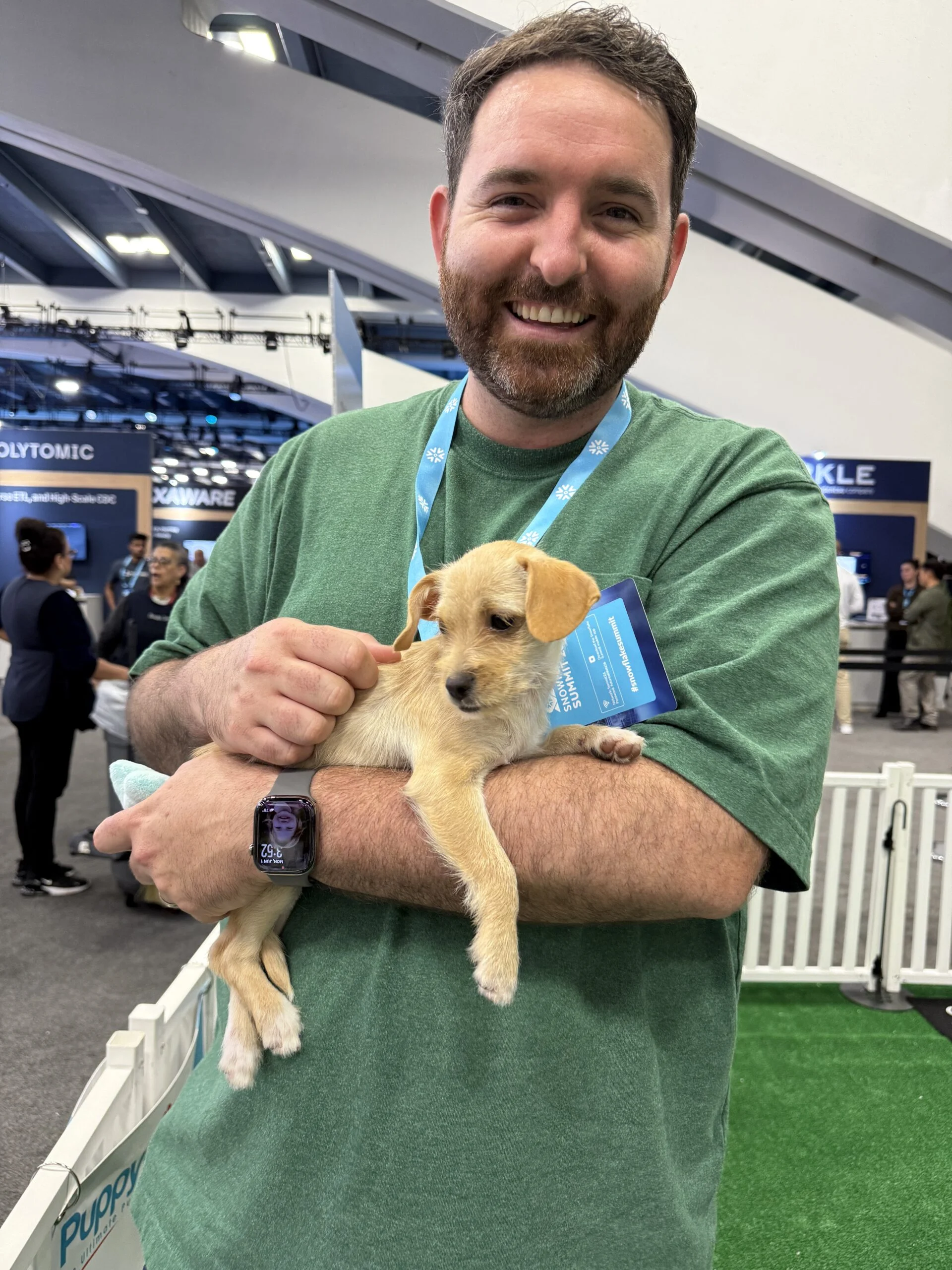 A smiling man with short brown hair and a beard, wearing a green t-shirt and an event lanyard, holds a small wire-haired light tan puppy in his arms.