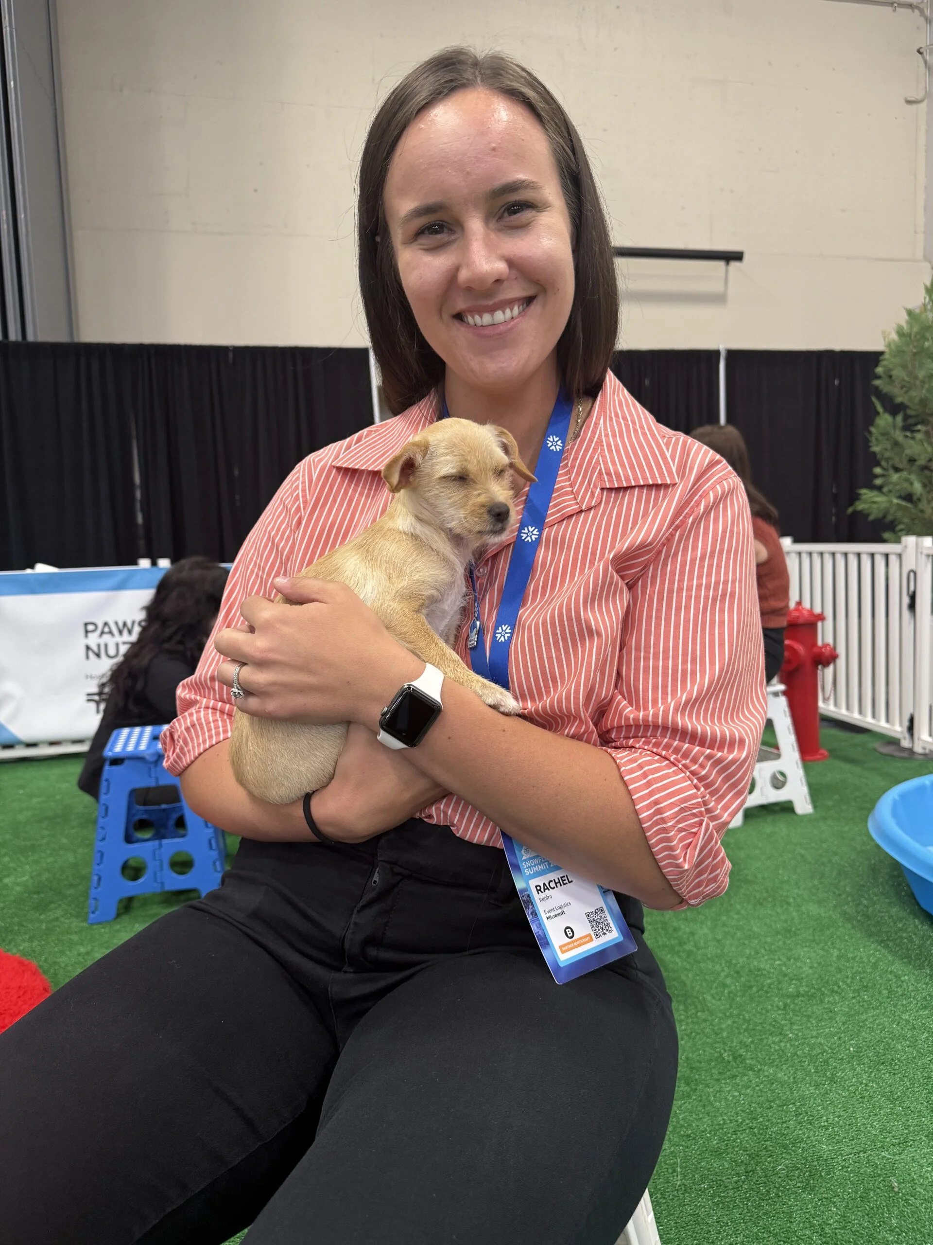A smiling woman named rachel with straight brown hair, wearing a coral striped button-up shirt and black pants, holds a small sleeping tan puppy in her arms.
