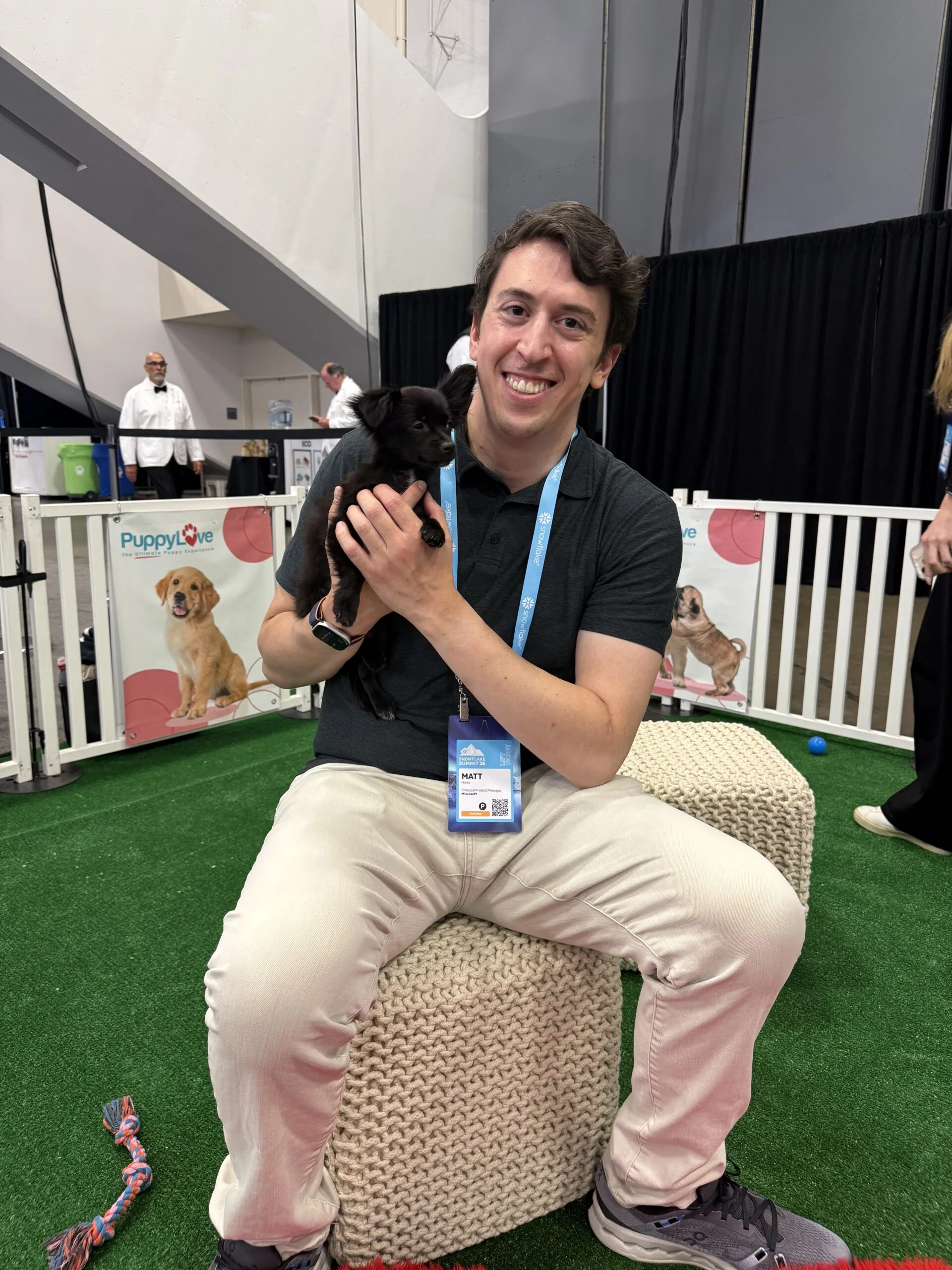 A smiling man named matt with short dark hair, wearing a dark grey polo shirt and light-colored pants, sits on a woven pouf while holding a tiny black puppy upright in his hands.