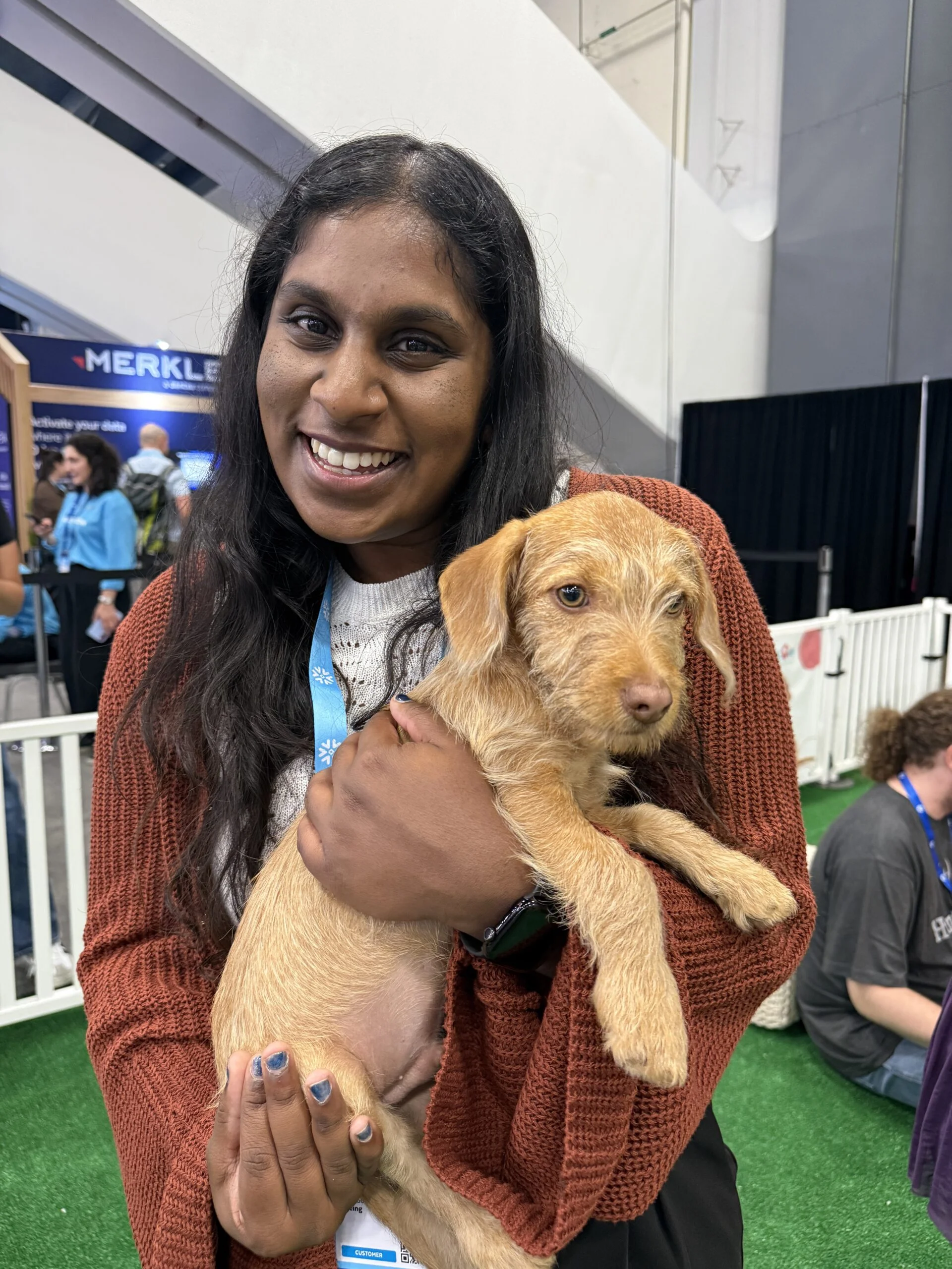 A smiling woman with long dark hair, wearing a rust-colored knit cardigan, holds a scruffy light tan puppy with floppy ears in her arms at an indoor event space.