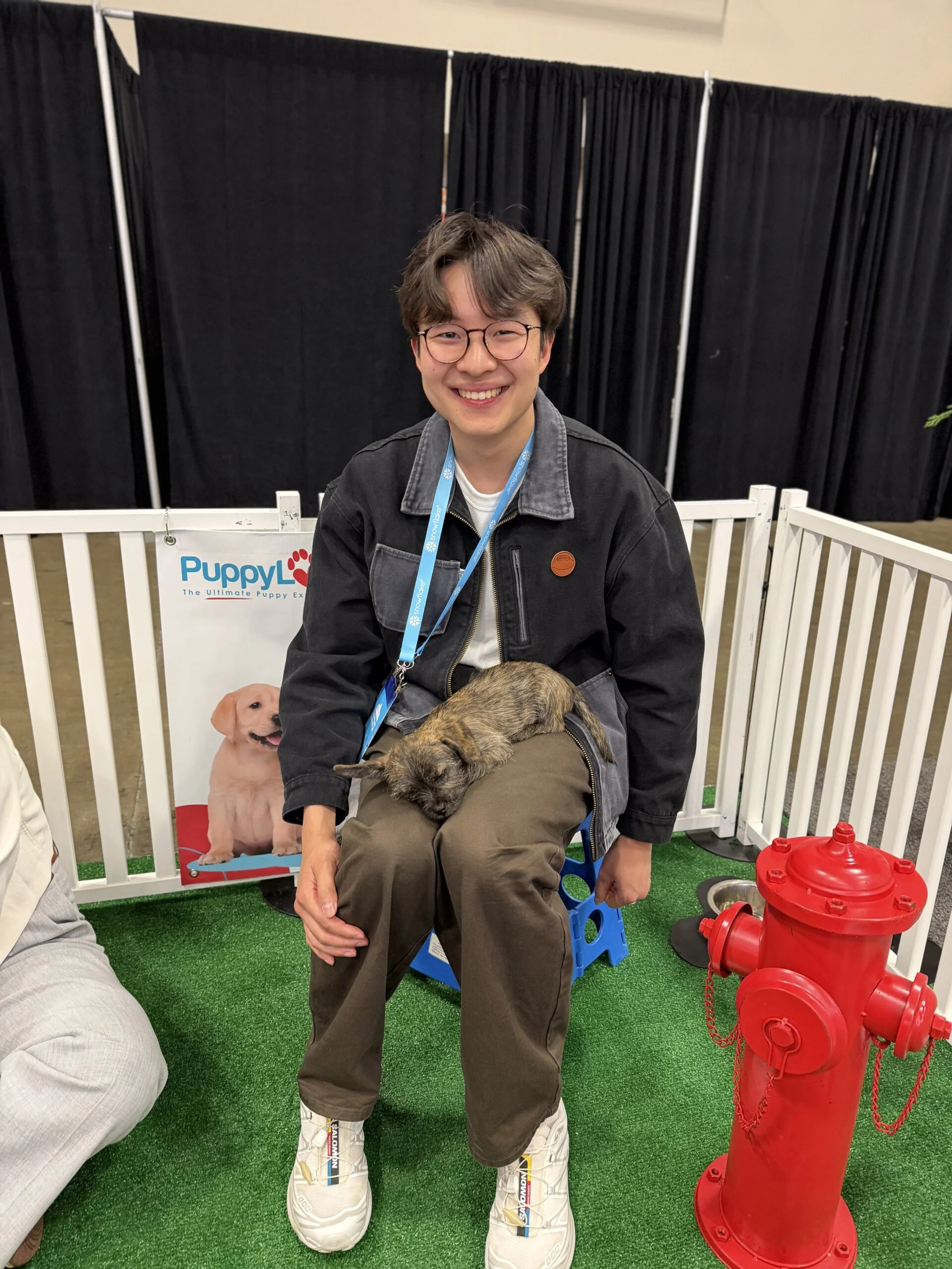 A smiling person with dark hair and round glasses, wearing a dark jacket and brown trousers, sits on a blue plastic stool while a small brindle puppy sleeps peacefully across their lap.