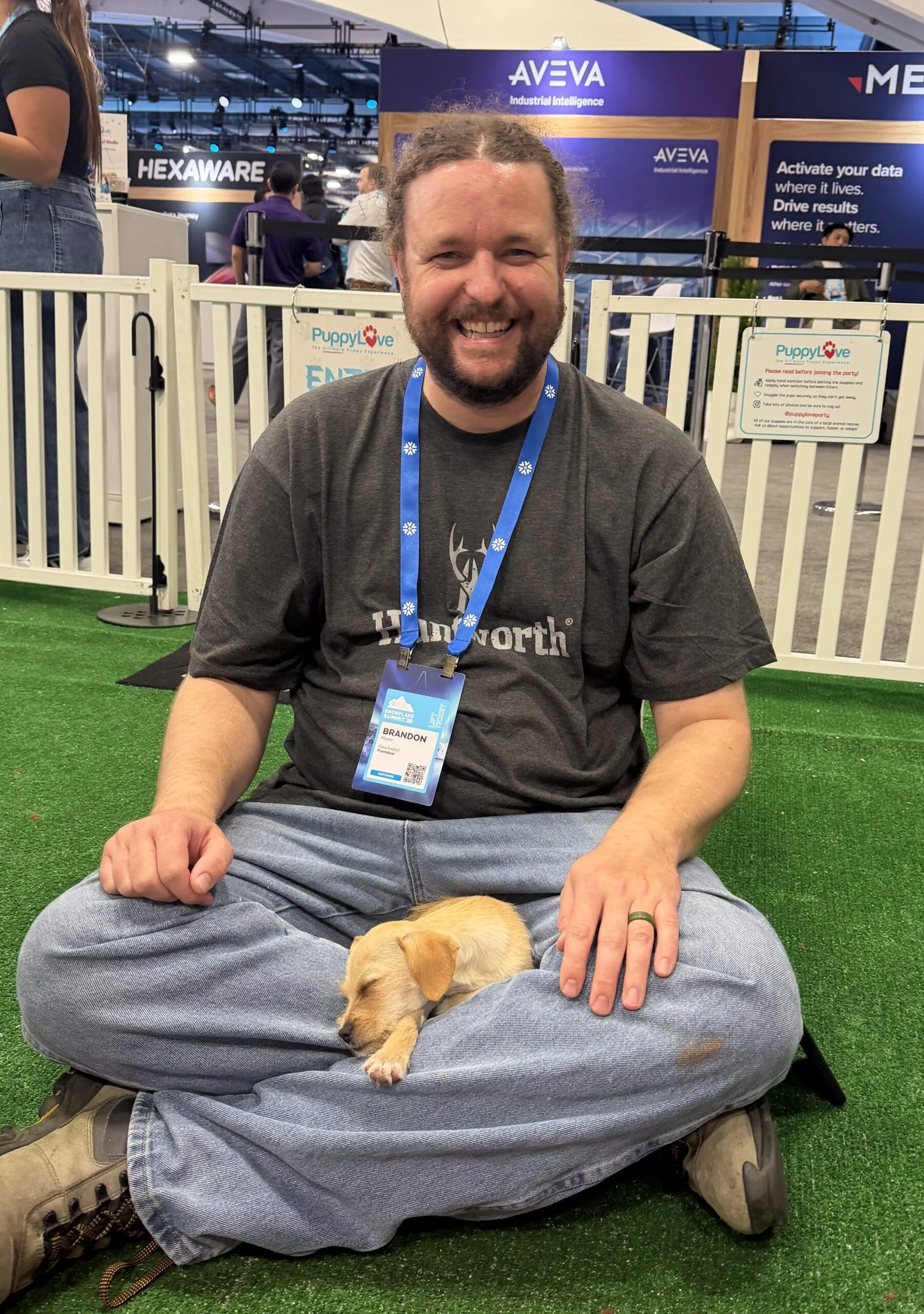 A smiling man named brandon with long hair tied back sits cross-legged on green artificial turf while a tiny, light tan puppy sleeps comfortably in his lap.
