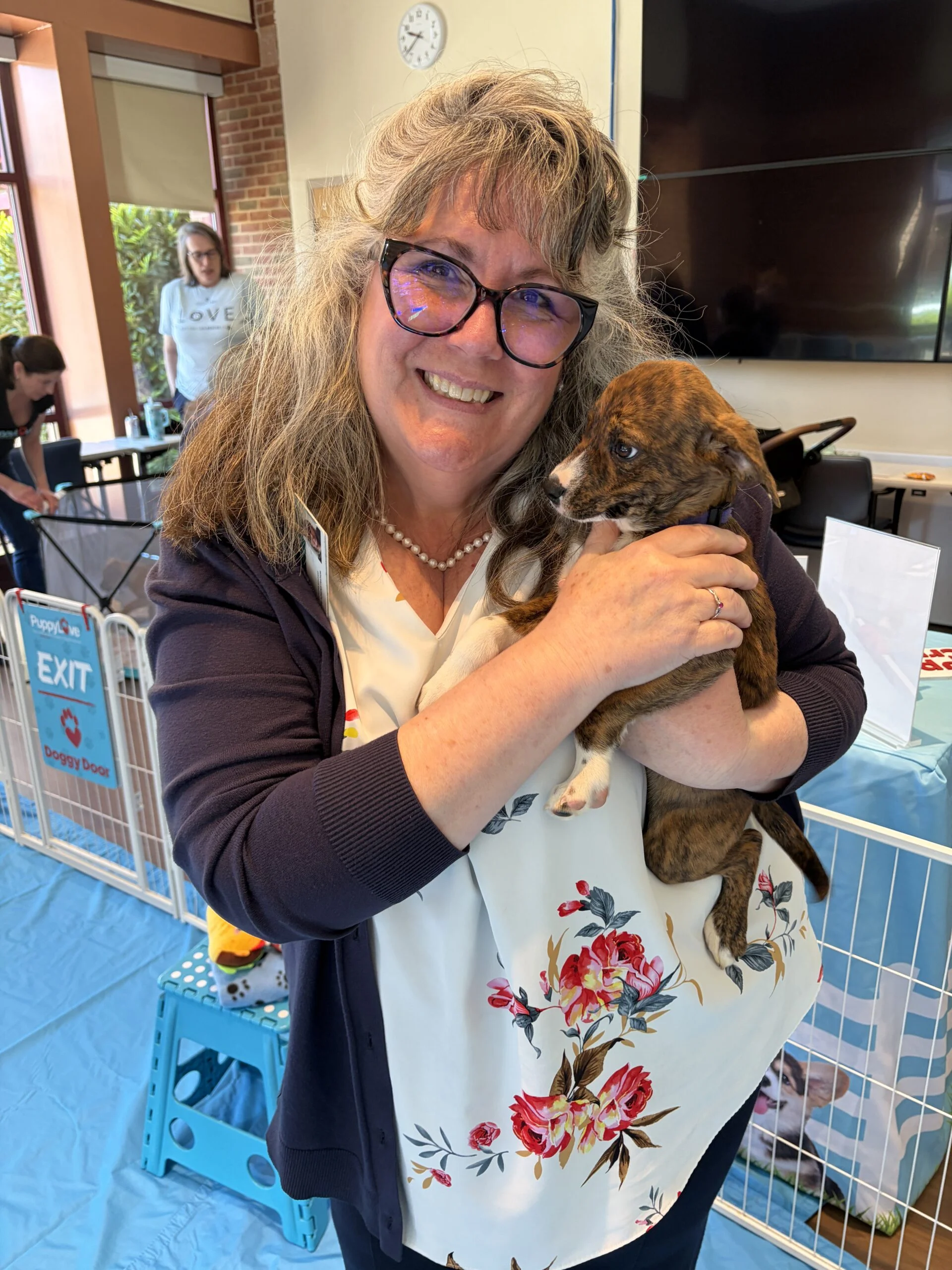 A smiling woman with glasses and a floral top holding a small brindle puppy at an event.