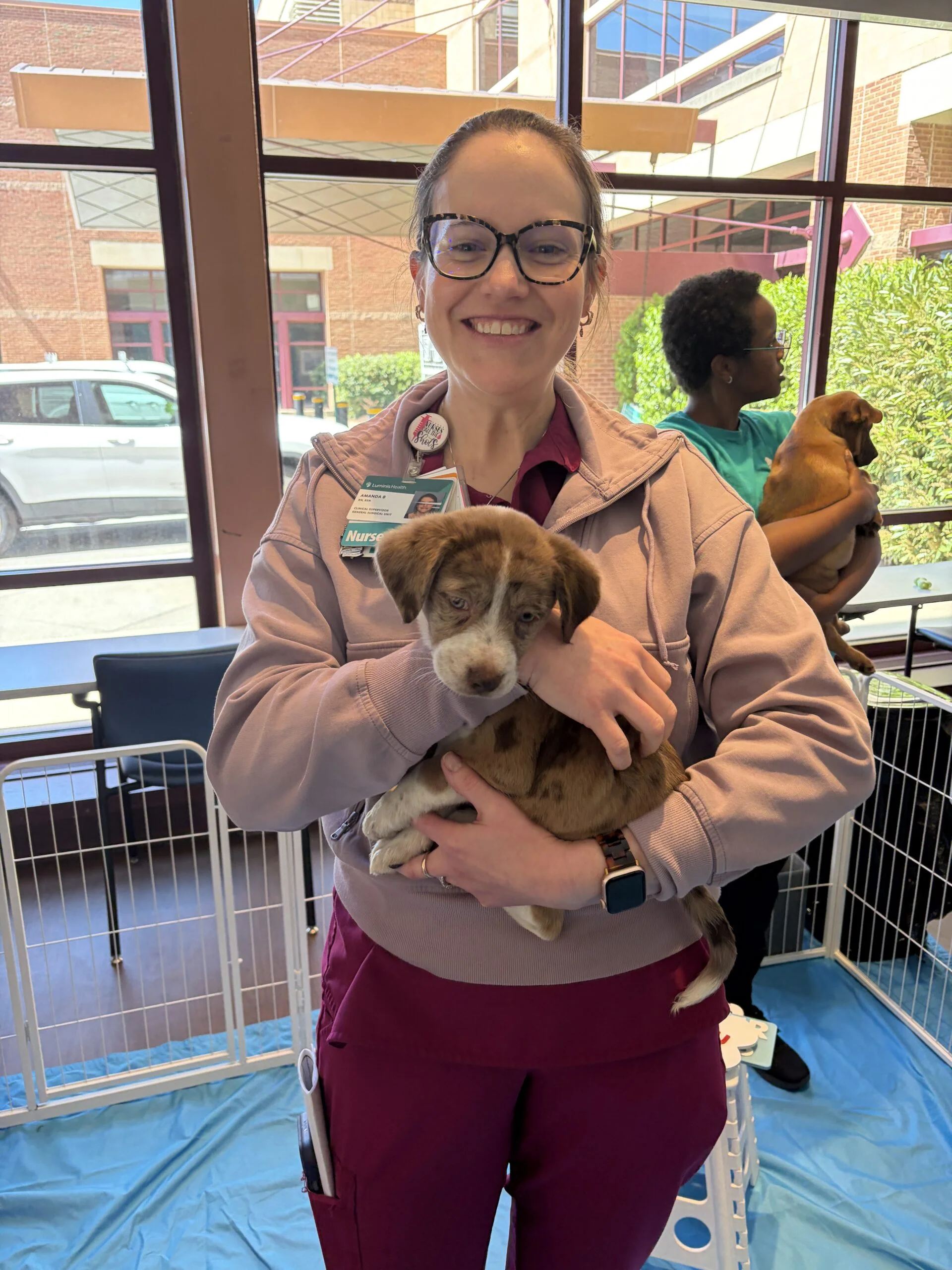 A smiling woman wearing glasses, a pink zip-up jacket, and maroon pants holding a small brown dappled puppy in a playpen.