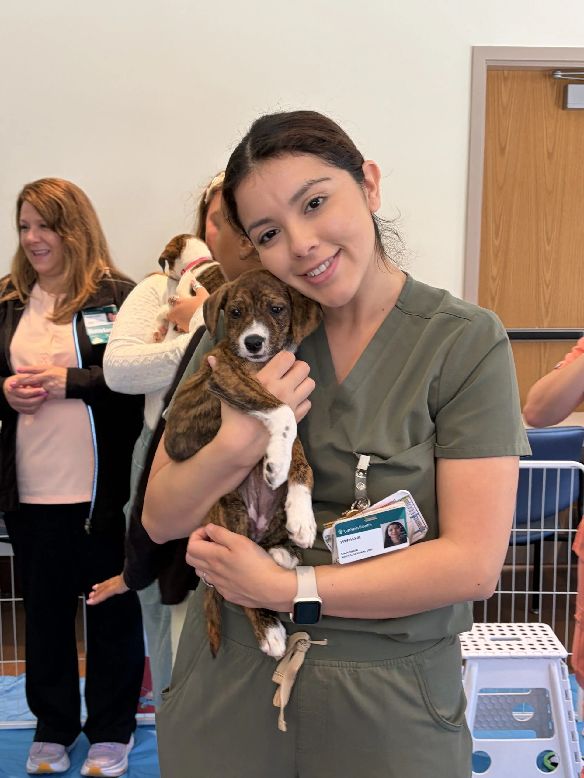 Healthcare worker in green scrubs smiling and holding a small brown and white puppy at luminis health aamc.