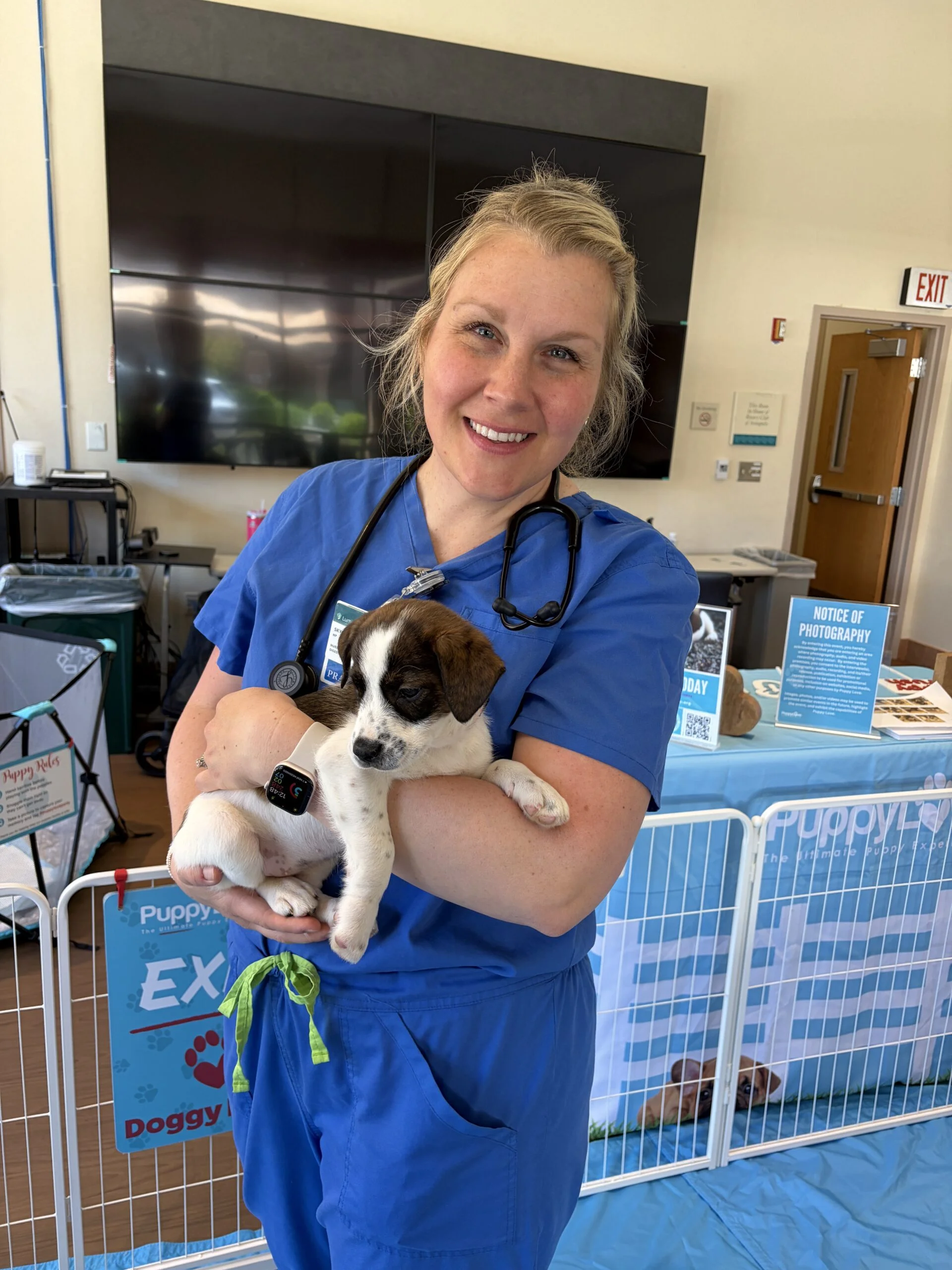 Healthcare worker in bright blue scrubs with a stethoscope smiling while holding a small brown and white puppy at luminis health aamc.