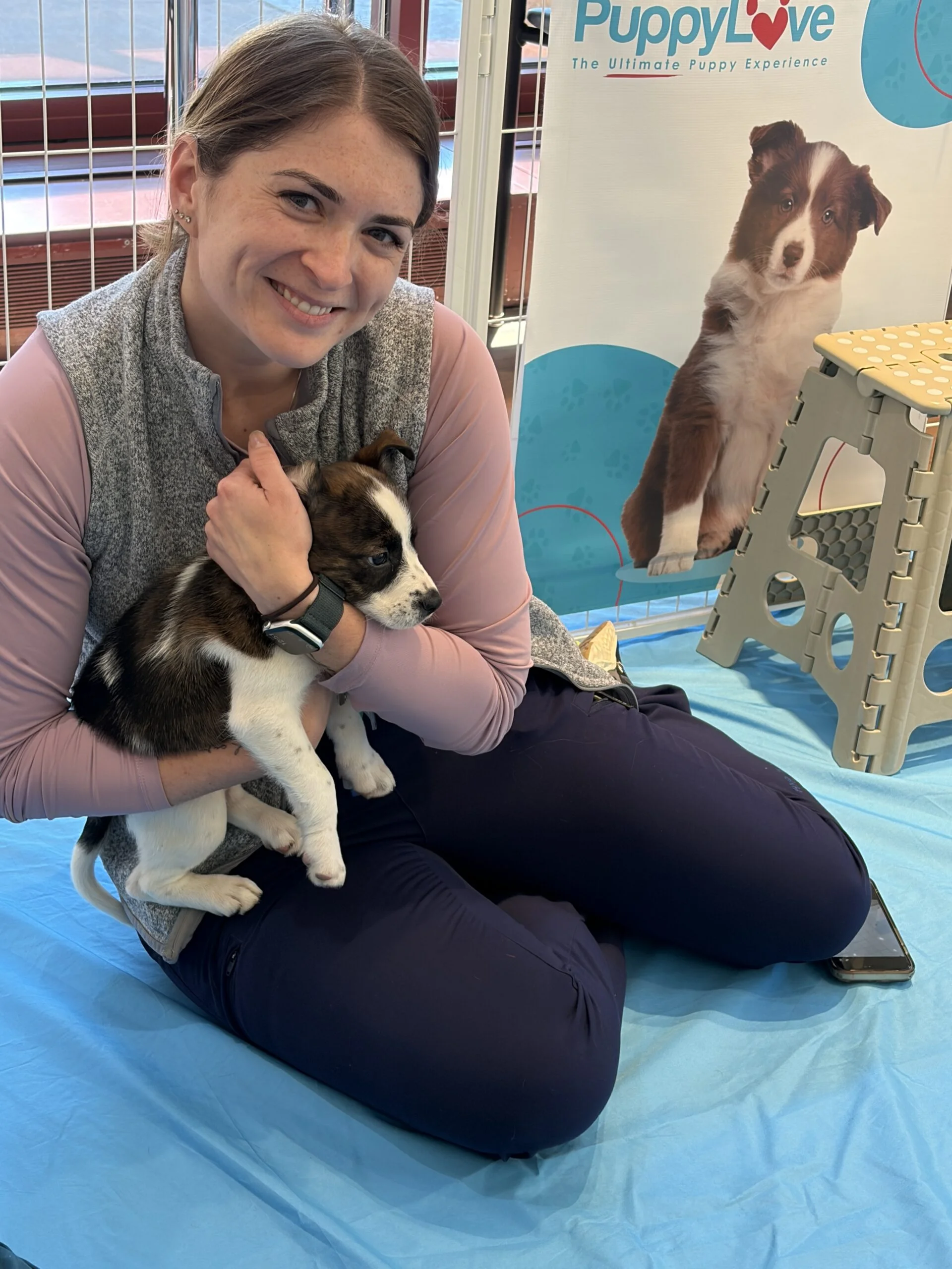 Woman in a grey vest kneeling and smiling while holding a black and white puppy at luminis health aamc.