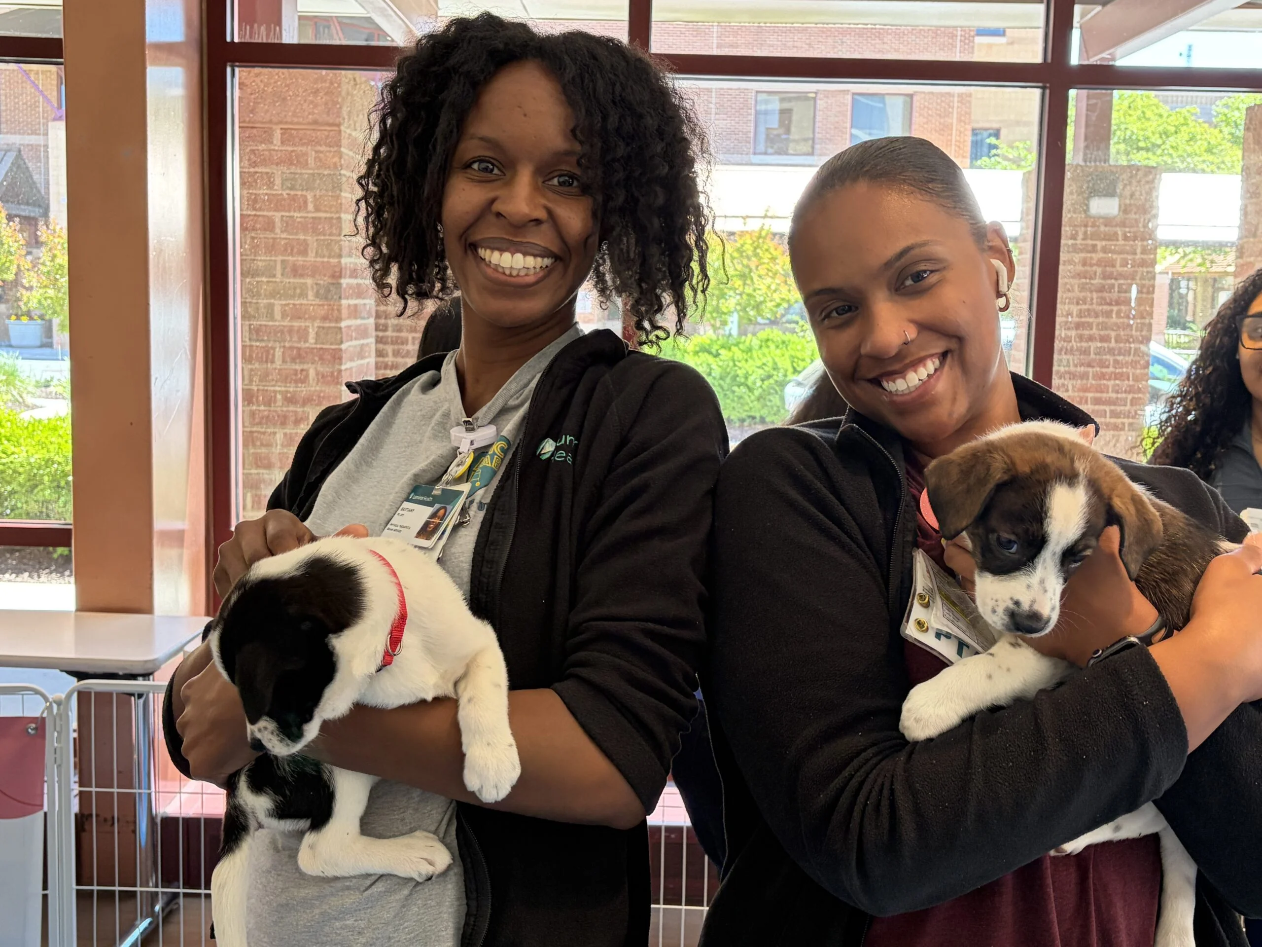Two healthcare workers smiling while holding a black-and-white puppy and a brown-and-white puppy at luminis health aamc.