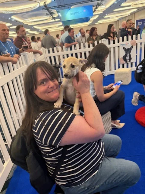 A smiling woman with dark hair and a navy-and-white striped shirt sitting on a blue carpeted floor while holding a small tan puppy with prominent ears.