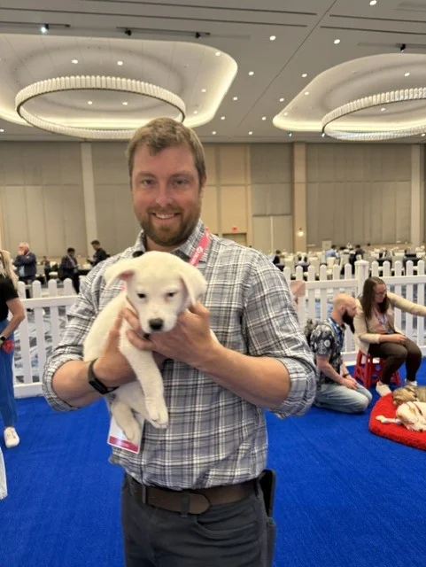 A smiling man with a beard and a plaid shirt stands holding a fluffy white puppy in his arms at an indoor event with blue carpeting.