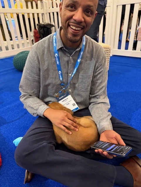 A smiling man wearing a grey button-down shirt sits cross-legged on a blue carpet with a small tan puppy sleeping in his lap while he holds a smartphone.