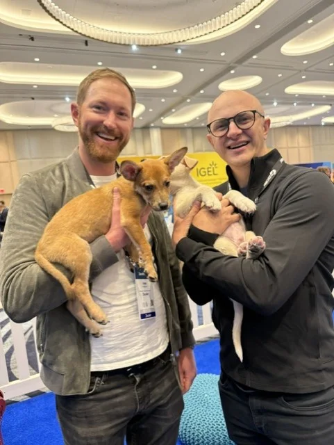 Two smiling men, one with a beard holding a tan puppy and one with glasses holding a white puppy, standing together inside a play area at a conference.