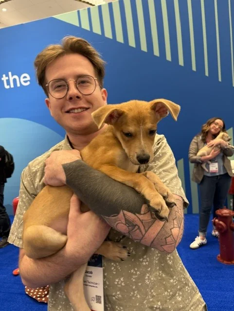 A smiling young man with glasses and a patterned shirt, featuring a prominent forearm tattoo, cradles a small tan puppy with alert ears in front of a blue backdrop.
