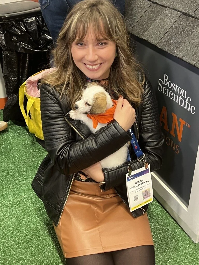 Smiling woman named hailey wearing a black leather jacket holding a small white and tan puppy with an orange bandana at an event.