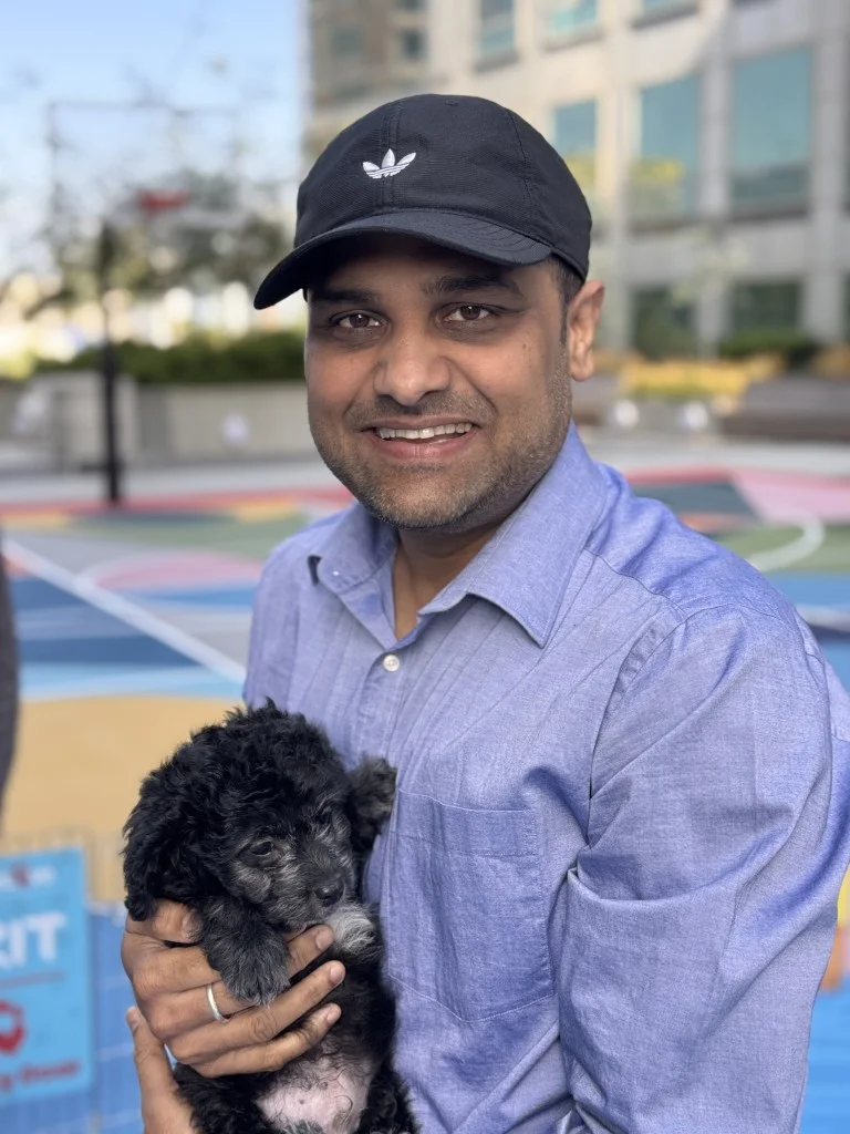 Smiling man wearing a black adidas baseball cap and light blue shirt holding a small fluffy black puppy outdoors.