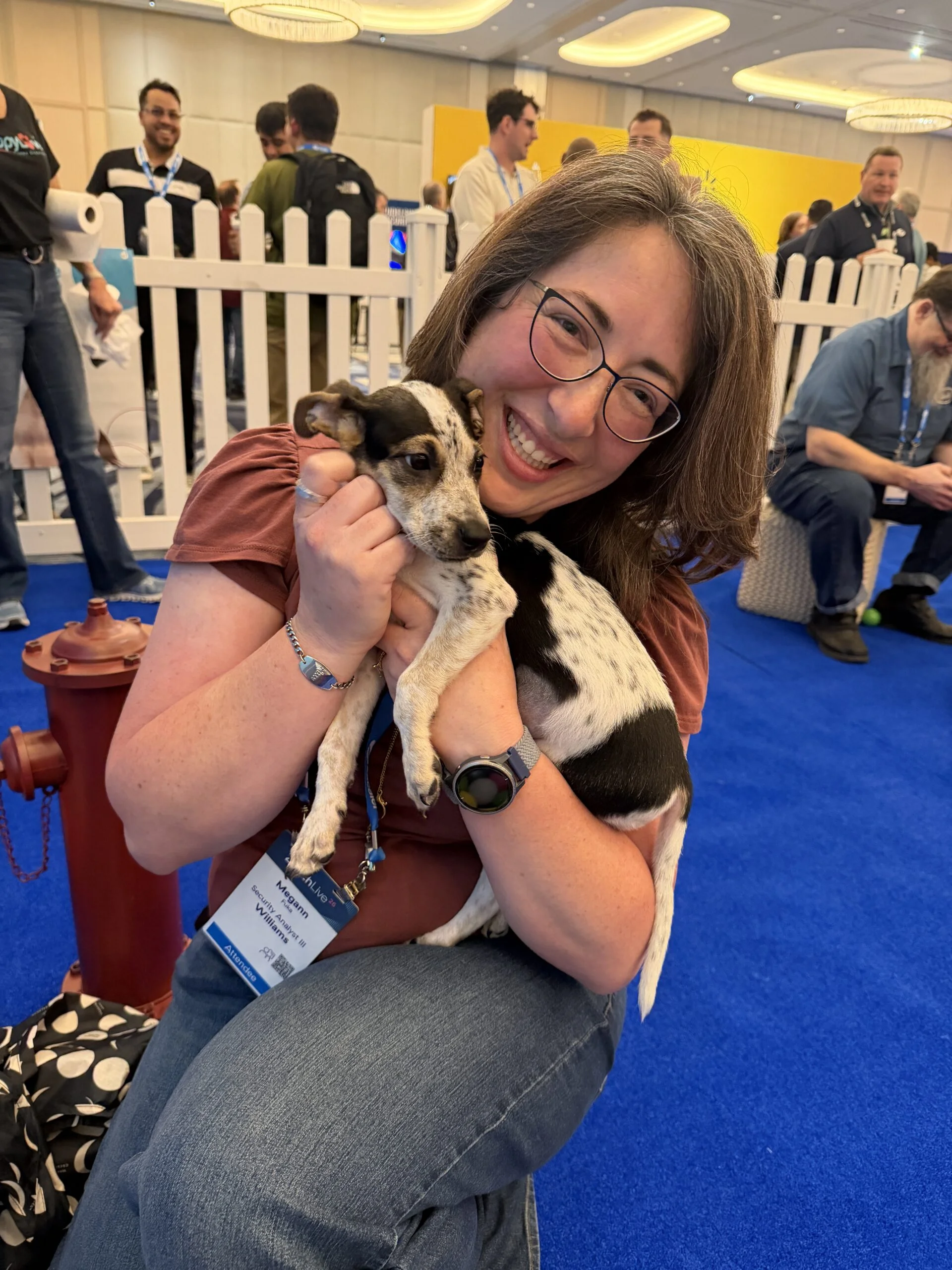 A smiling woman with glasses and brown hair wearing a mauve shirt sitting on a blue carpeted floor while cradling a small speckled black-and-white puppy.
