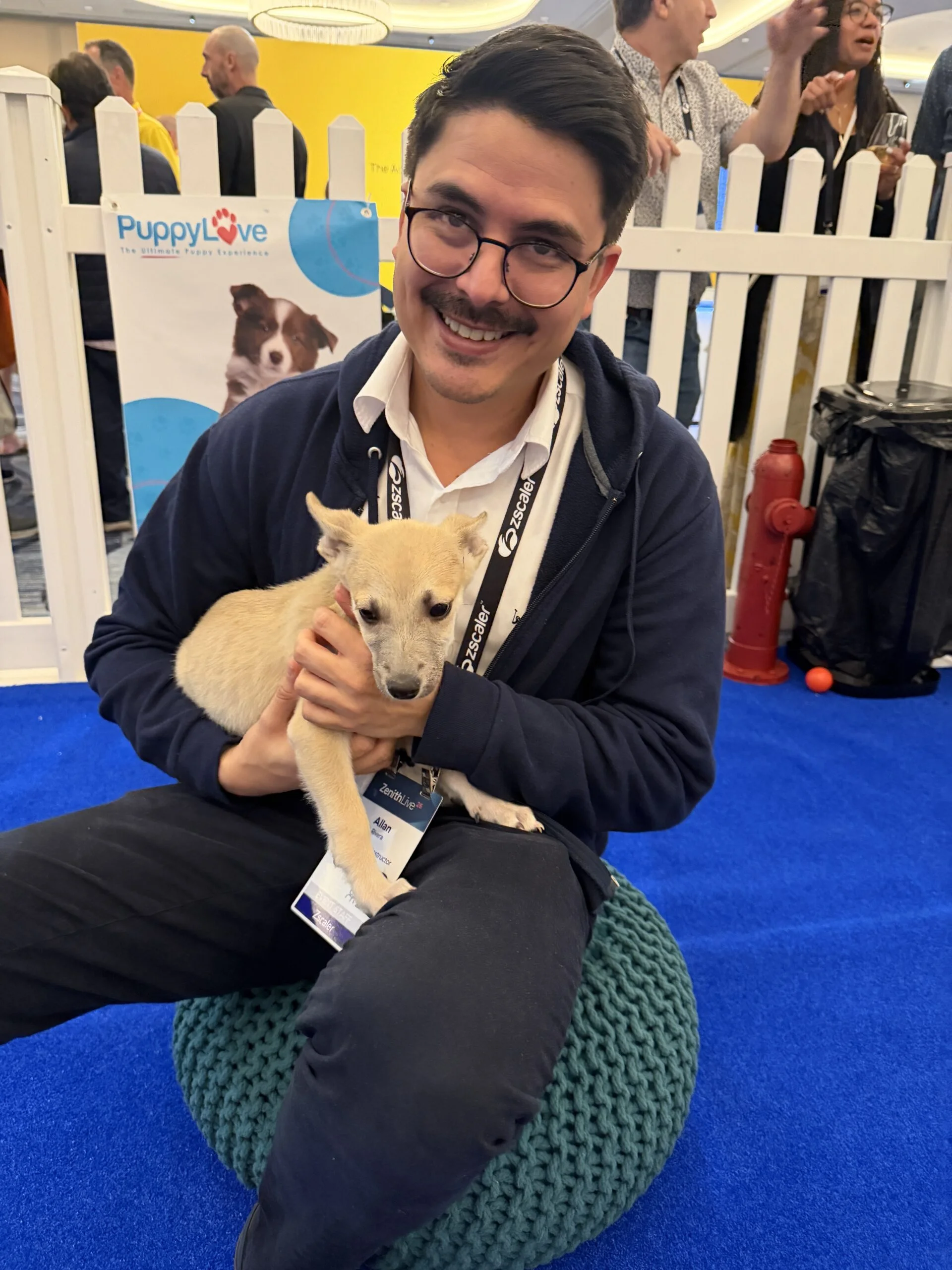 A smiling man with glasses sits on a teal knit pouf on a blue carpet, holding a small tan puppy.