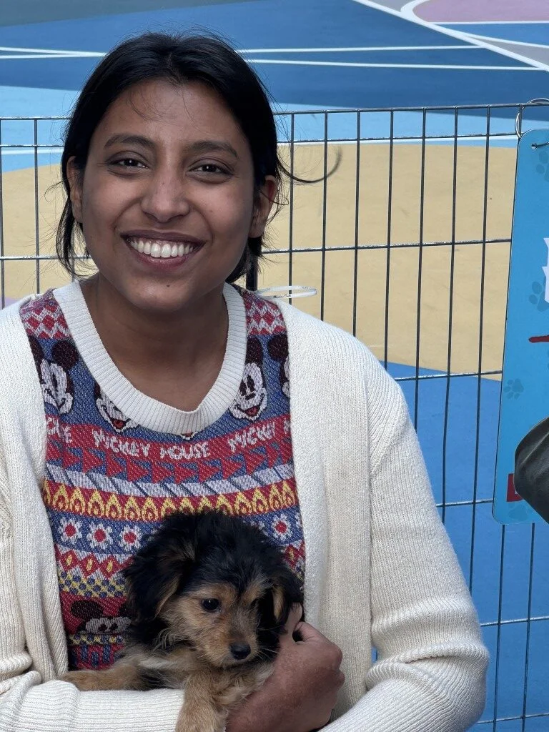 Smiling woman with dark hair wearing a mickey mouse sweater holding a small scruffy tan puppy.