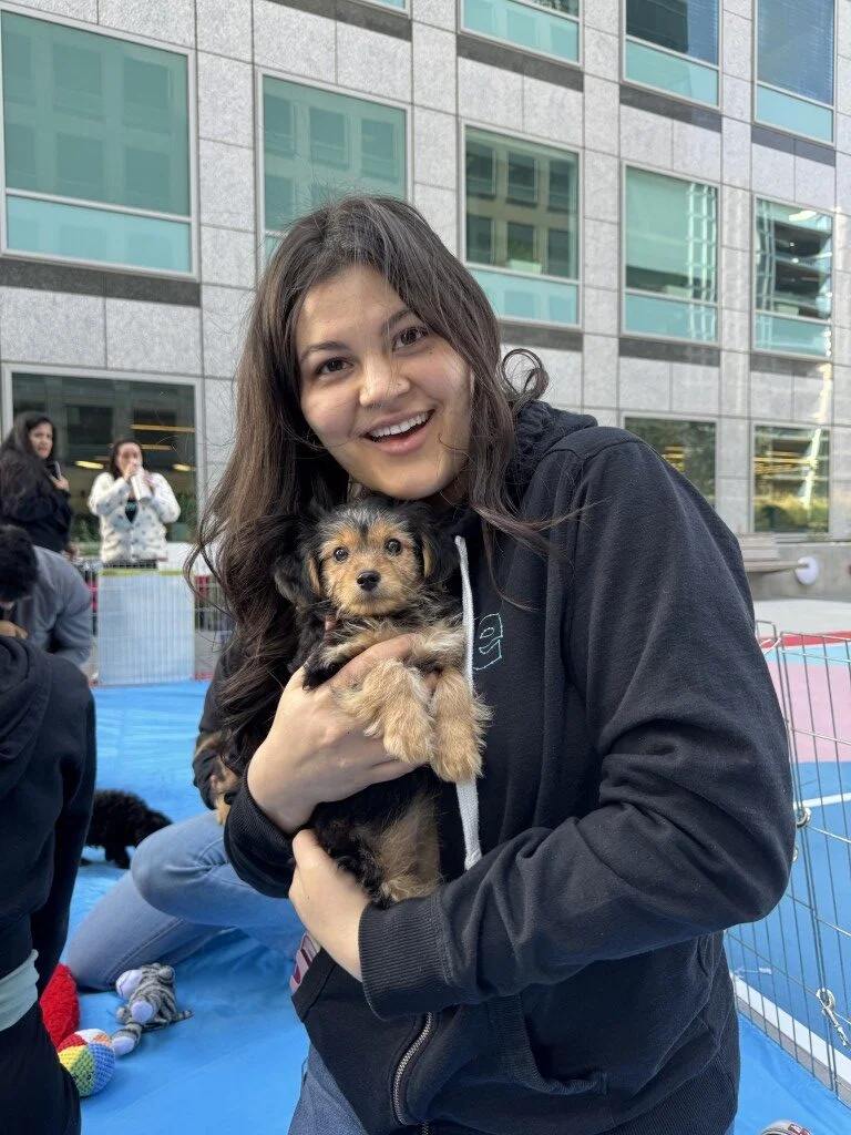 Smiling woman in a dark hoodie holding a small scruffy tan and black puppy outdoors.