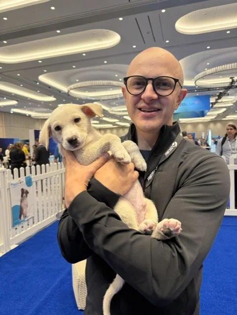 A smiling bald man with black glasses and a dark zip-up jacket cradling a small white puppy inside an indoor event play area.