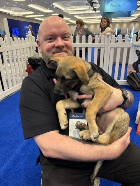 A smiling bald man with a beard, wearing a black polo shirt, cradles a tan puppy with a dark muzzle inside an indoor conference play area.