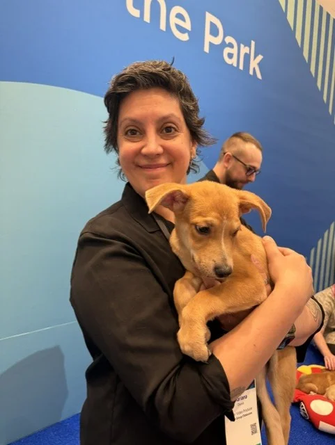 A smiling person with short, wavy dark hair wearing a black shirt cradles a small tan puppy with floppy ears in front of a blue background.